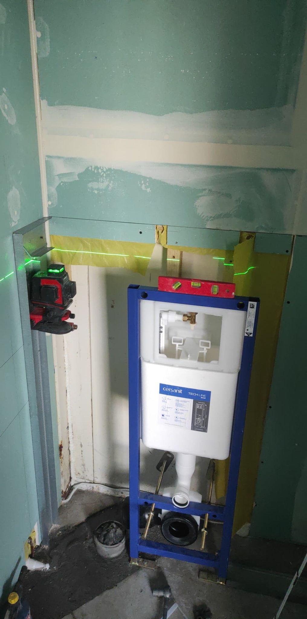 Stelaż podtynkowy Cersanit zamontowany w narożniku łazienki, widoczny laser krzyżowy, ściany częściowo obłożone płytą kartonowo-gipsową, poziomica na stelażu.