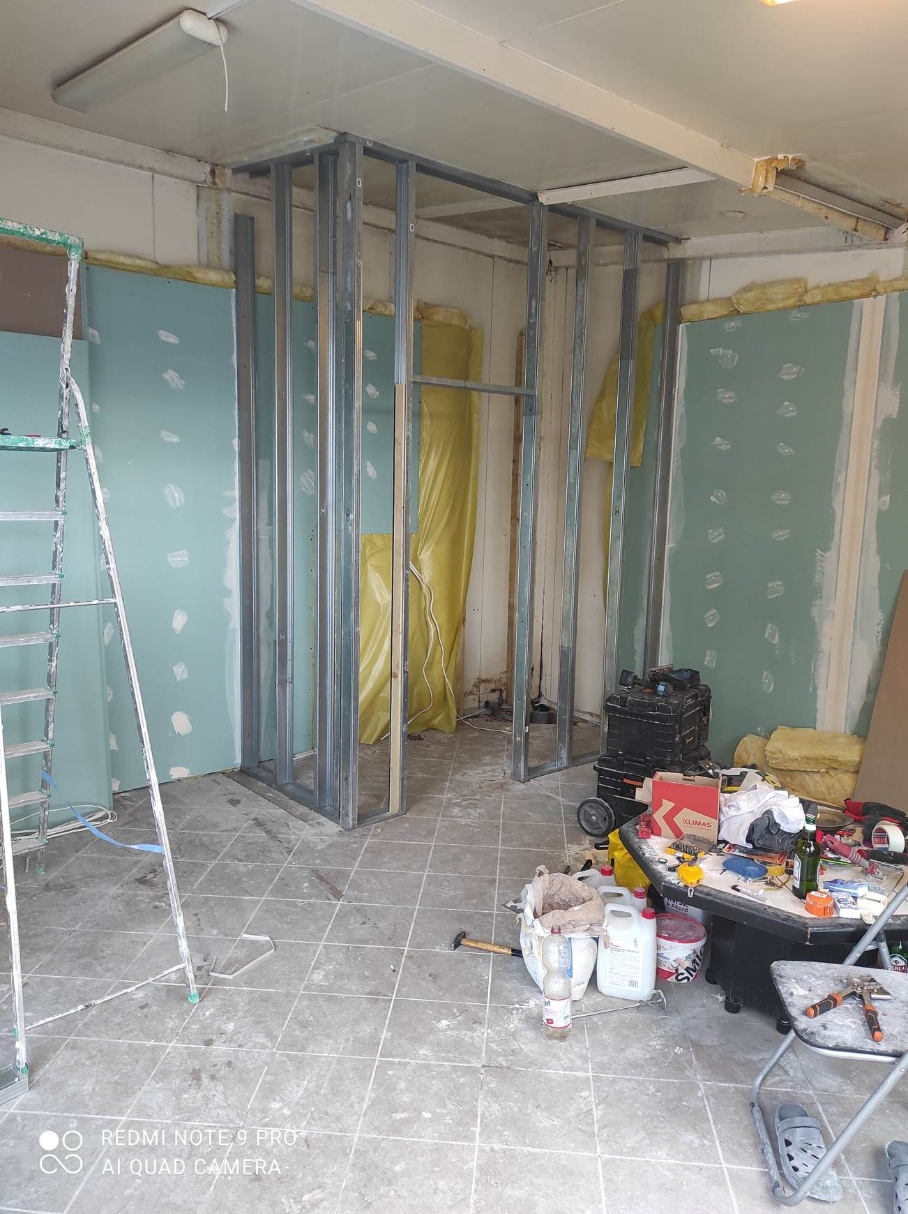 Wnętrze w trakcie prac wykończeniowych: metalowy szkielet ścian działowych, częściowo obłożony zieloną płytą, widoczna izolacja z żółtej wełny mineralnej, narzędzia i materiały budowlane...