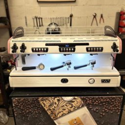 CoffeeRiver - Profesjonalny ekspres do kawy z trzema kolbami, podświetlony, stoi na szafce z motywem kawy i paczką kawy Filicori, w tle widoczne narzędzia.