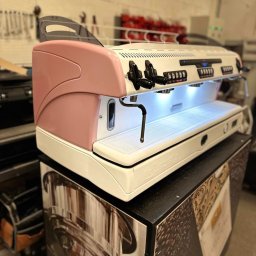 CoffeeRiver - Profesjonalny ekspres do kawy La Spaziale w kolorze białym z różowymi akcentami, umieszczony na szafce z grafiką kawy, widoczne przyciski i dysze, oświetlenie LED w strefie filiżanek.