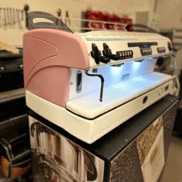 Profesjonalny ekspres do kawy La Spaziale w kolorze białym z różowymi akcentami, umieszczony na szafce z grafiką kawy, widoczne przyciski i dysze, oświetlenie LED w strefie filiżanek.