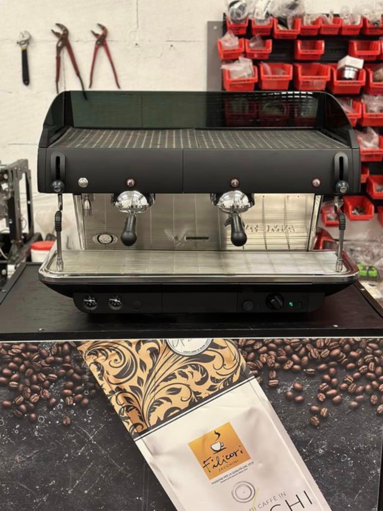 Profesjonalny, dwugrupowy ekspres do kawy w kolorze czarnym i srebrnym, z opakowaniem kawy Filicori Zecchini na pierwszym planie, widoczne narzędzia i pojemniki w tle.