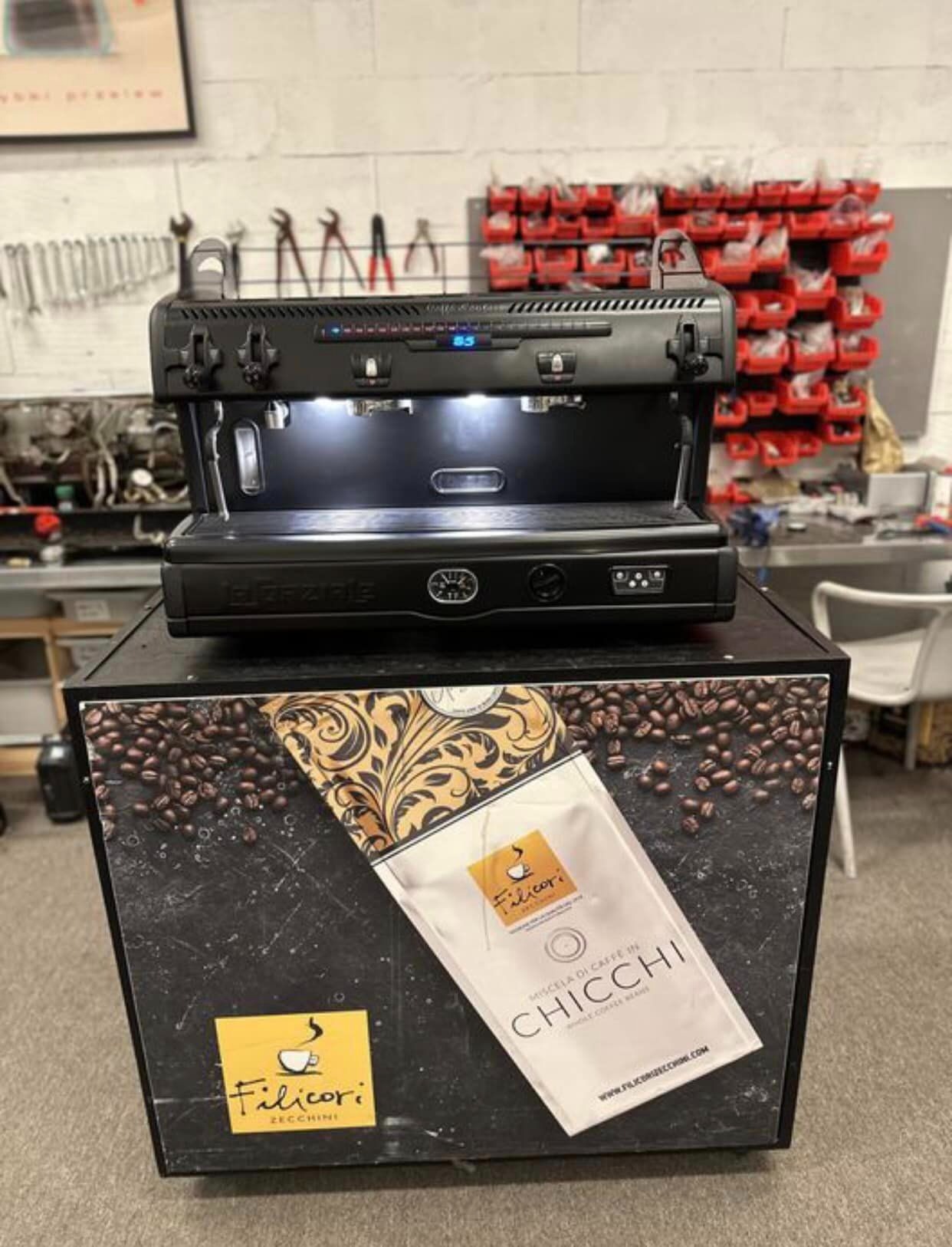 Profesjonalny ekspres do kawy La Spaziale na czarnym postumencie z grafiką kawy i logo Filicori Zecchini, narzędzia warsztatowe w tle.