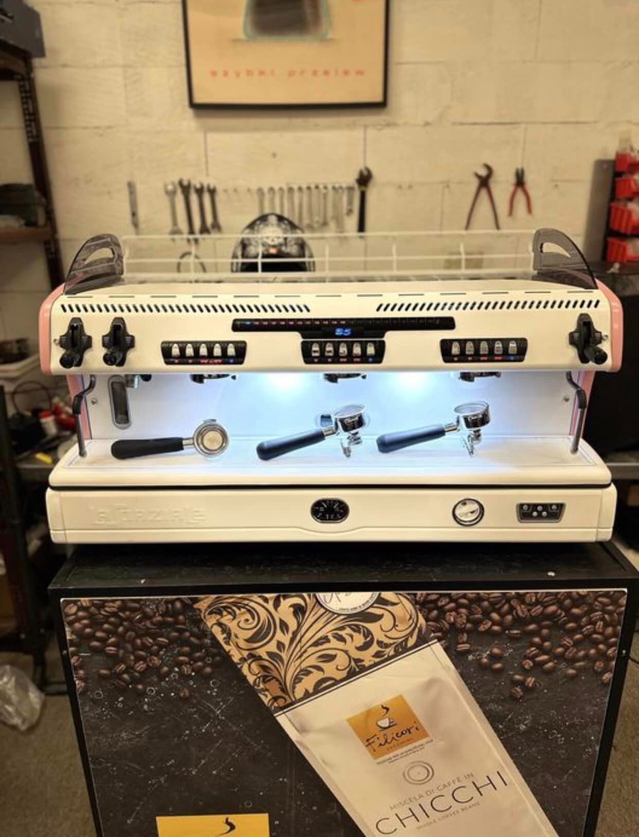 Profesjonalny ekspres do kawy z trzema kolbami, podświetlony, stoi na szafce z motywem kawy i paczką kawy Filicori, w tle widoczne narzędzia.