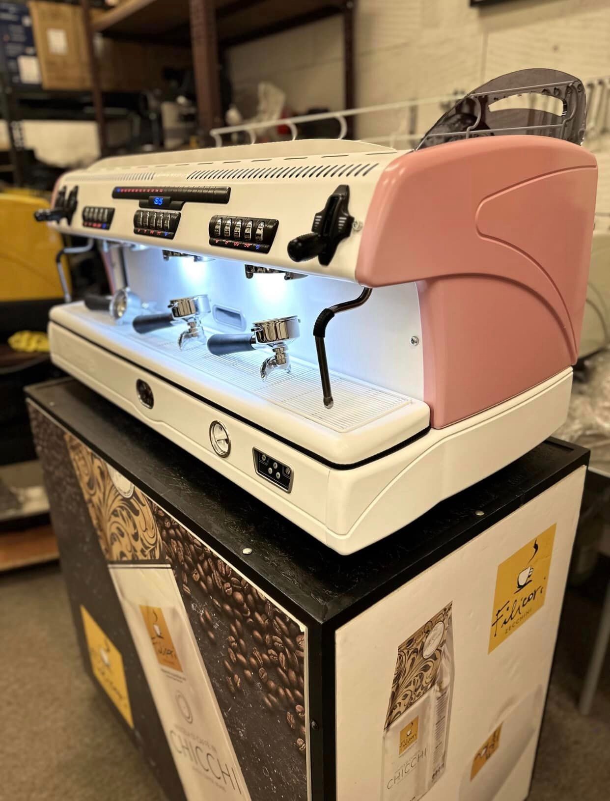 Profesjonalny, dwugrupowy ekspres do kawy w kolorze białym z różowymi akcentami, umieszczony na czarnym postumencie z grafiką ziaren kawy i logo marki Chicchi.