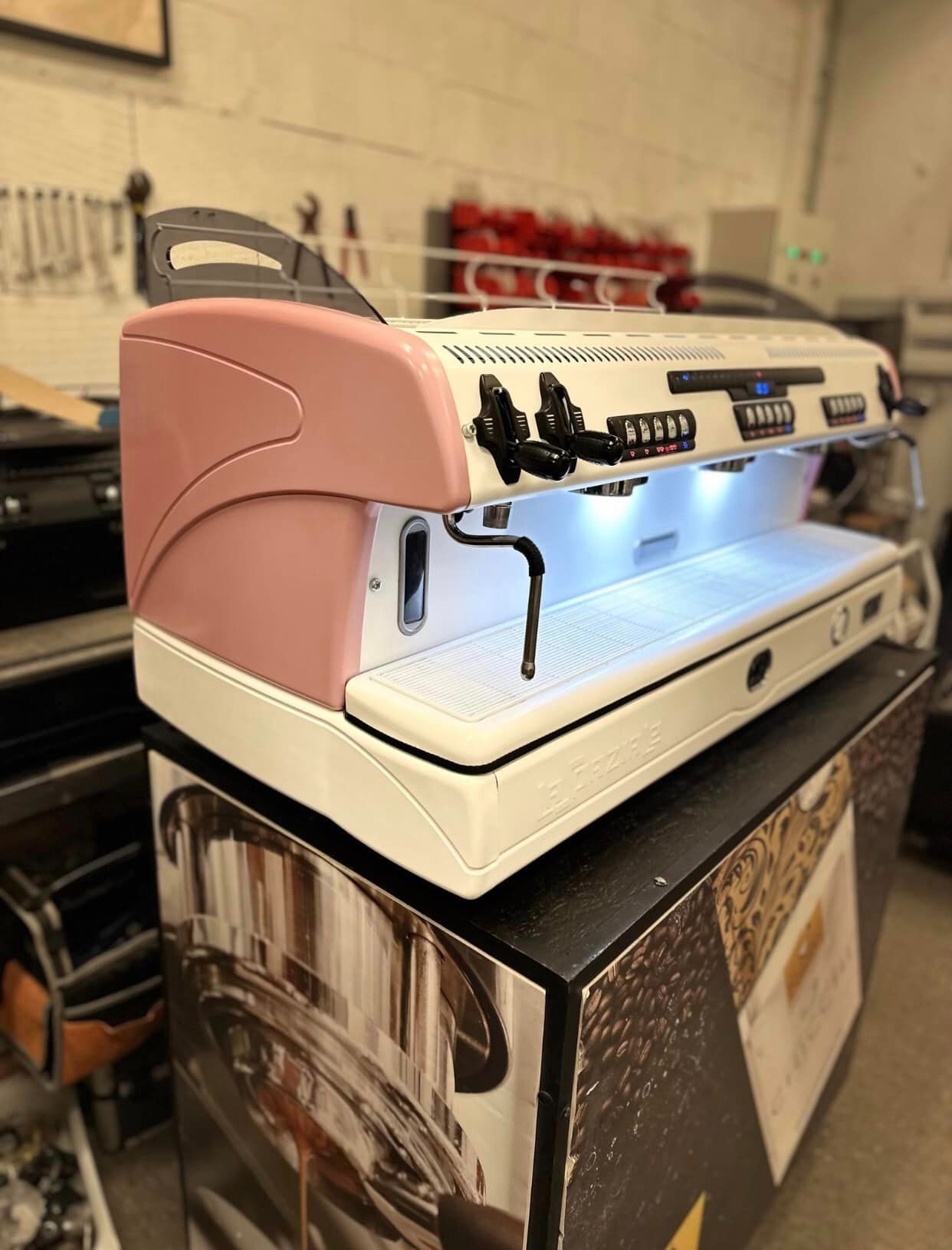 Profesjonalny ekspres do kawy La Spaziale w kolorze białym z różowymi akcentami, umieszczony na szafce z grafiką kawy, widoczne przyciski i dysze, oświetlenie LED w strefie filiżanek.