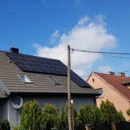 Dom z panelami słonecznymi na dachu, widok z dołu, błękitne niebo z chmurami w tle, antena satelitarna na ścianie, tuje w ogrodzie.