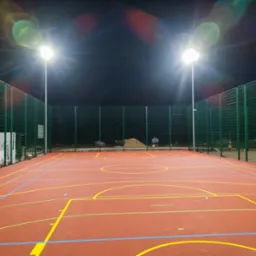 Oświetlony nocą wielofunkcyjny plac zabaw z czerwoną nawierzchnią, zielonym ogrodzeniem i widocznymi liniami do różnych gier zespołowych.