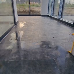 Remonty de lux - Nowa, ciemna, błyszcząca posadzka z płytek w przestronnym wnętrzu z dużymi oknami, widoczne refleksy światła i ślady po myciu.