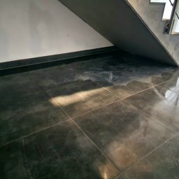 Remonty de lux - Błyszcząca, ciemnoszara podłoga z płytek ceramicznych pod schodami, widoczny fragment ściany z listwą przypodłogową.