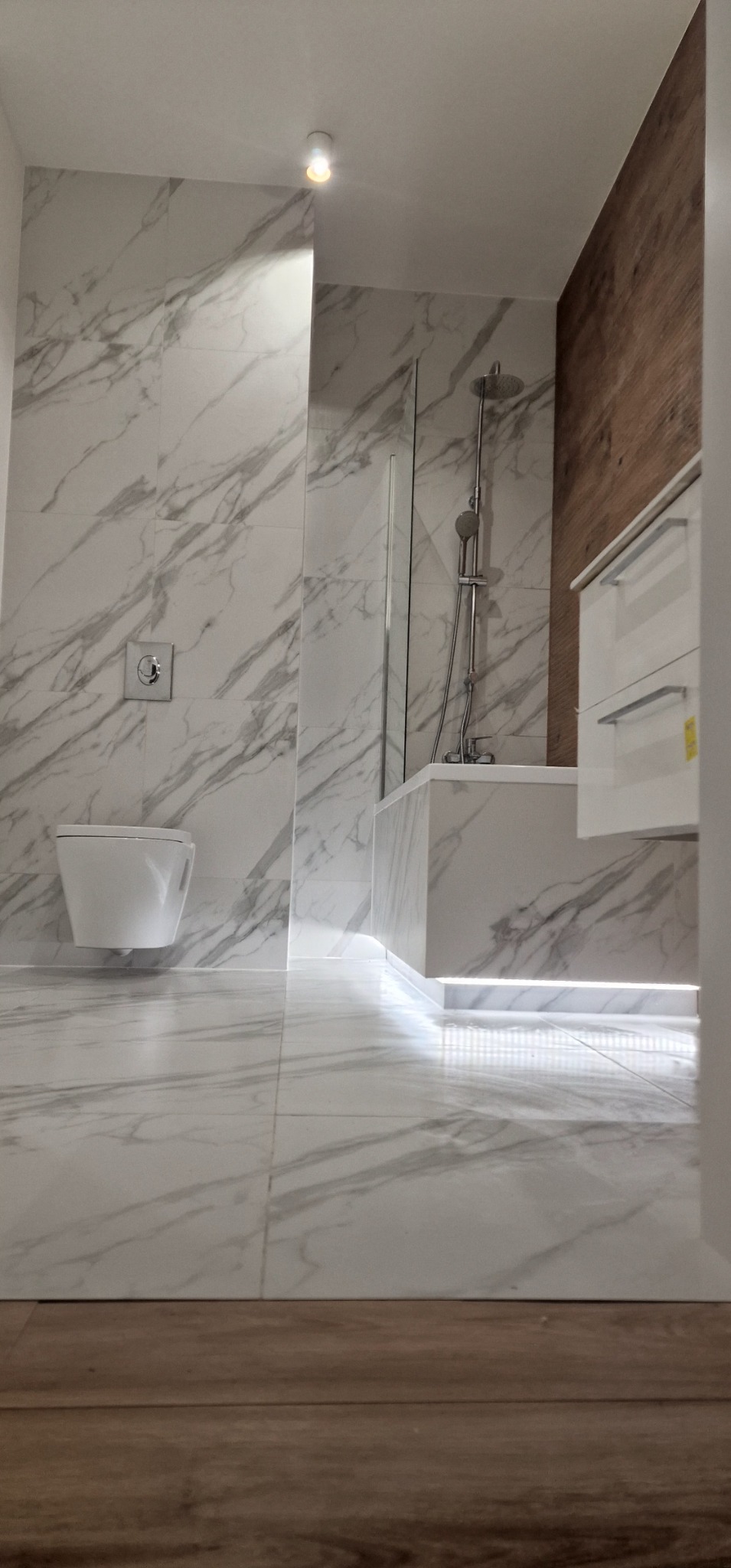 Wykończona łazienka z białymi marmurkowymi płytkami na ścianach i podłodze, podświetlana wanna, wisząca toaleta i drewniana ściana z szafką.