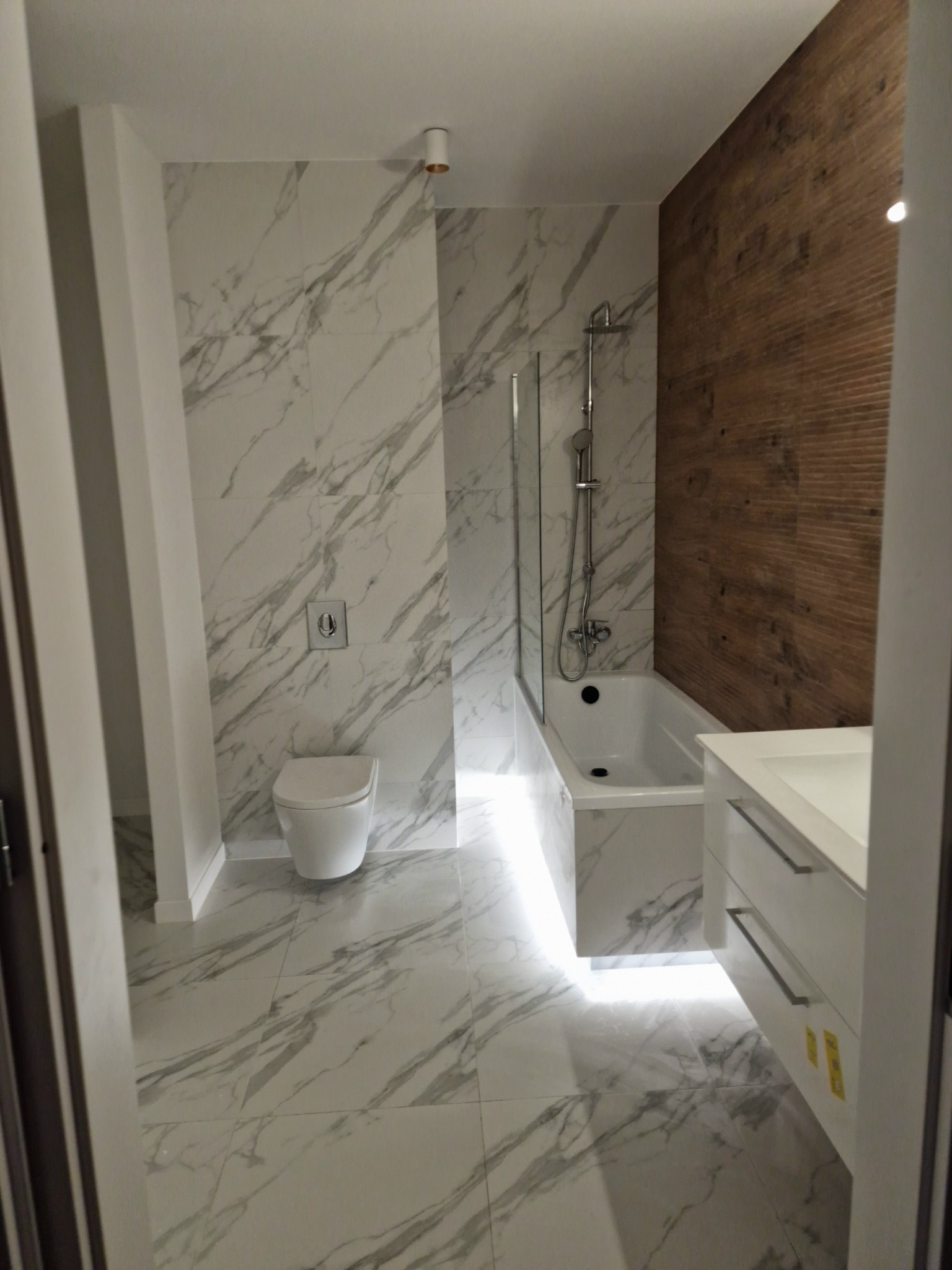 Nowoczesna łazienka z białymi marmurkowymi płytkami, drewnopodobną ścianą, podświetlaną wanną i wiszącą toaletą z chromowanym przyciskiem.