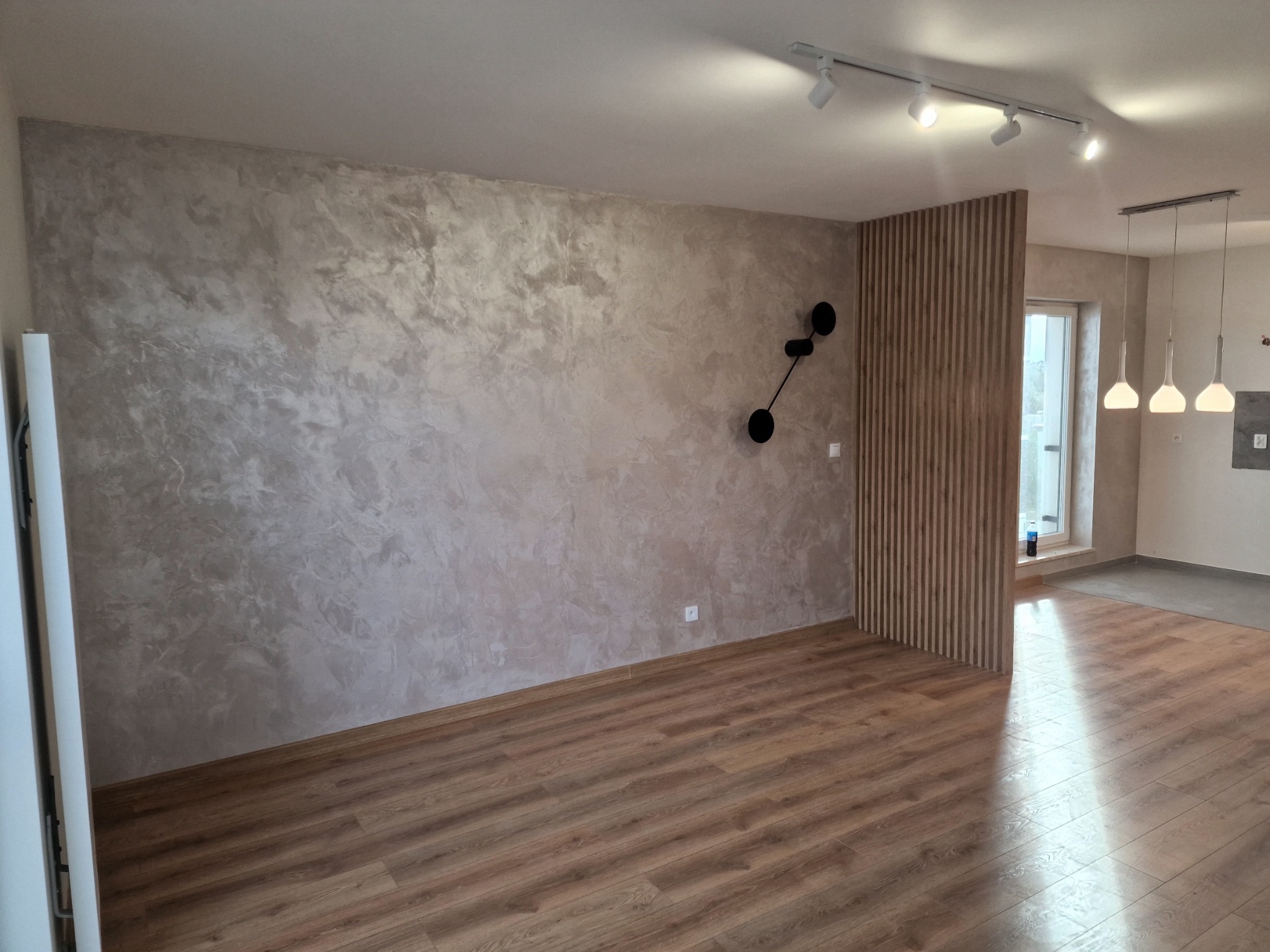 Wykończone wnętrze: ściana z dekoracyjnym tynkiem w odcieniach beżu i szarości, drewniana podłoga, lampy sufitowe oraz dekoracyjny panel z pionowych listew.