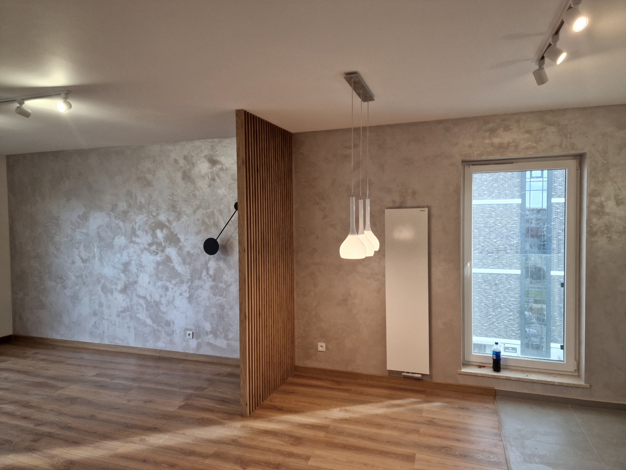 Wnętrze pokoju z drewnianą podłogą, ścianami pokrytymi dekoracyjnym tynkiem w odcieniach szarości, drewnianą ażurową ścianką działową, nowoczesną lampą wiszącą i grzejnikiem panelowym.