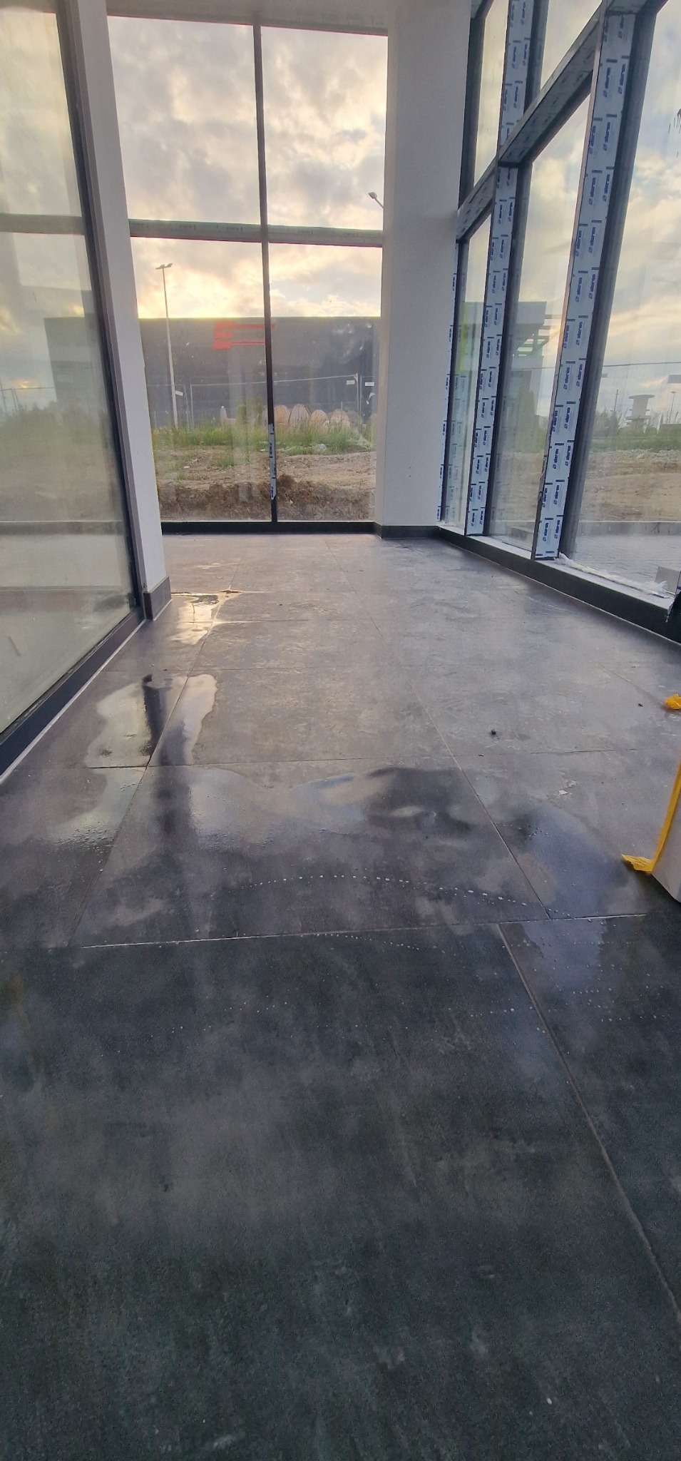 Nowa, ciemna, błyszcząca posadzka z płytek w przestronnym wnętrzu z dużymi oknami, widoczne refleksy światła i ślady po myciu.