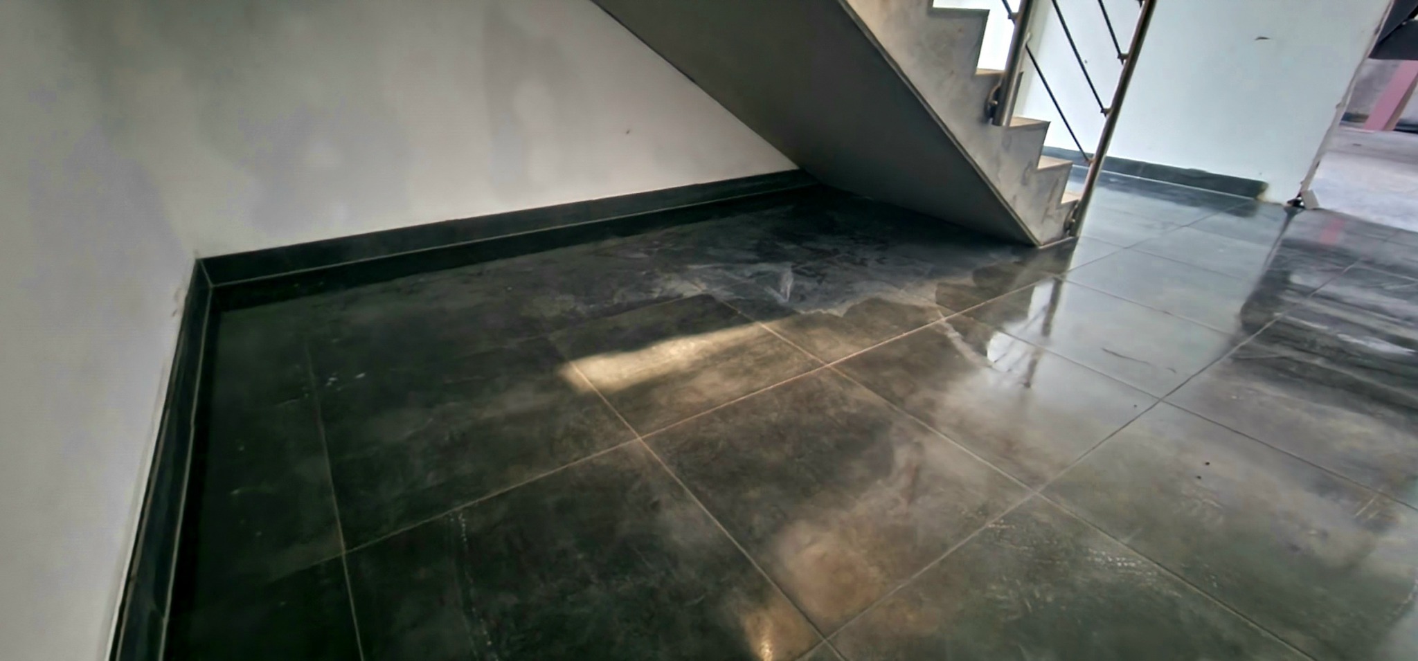 Błyszcząca, ciemnoszara podłoga z płytek ceramicznych pod schodami, widoczny fragment ściany z listwą przypodłogową.