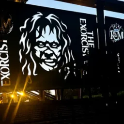 Nowoczesna balustrada z motywami filmowymi i logo firmy, wykonana z metalu, podświetlona od dołu, widok z dołu na tle wieczornego nieba.