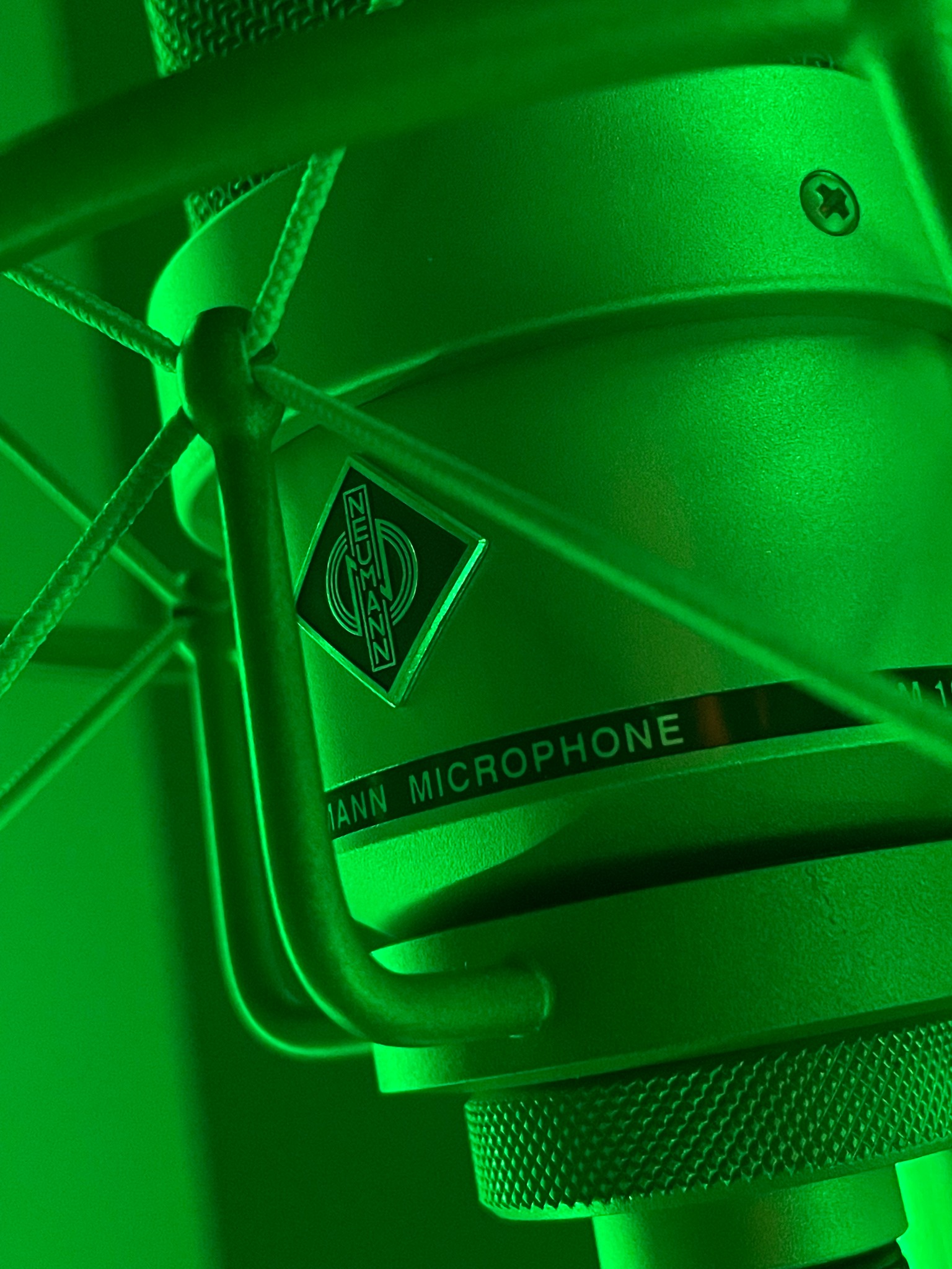 Kondensatorowy mikrofon studyjny Neumann w uchwycie antywibracyjnym, oświetlony intensywnym zielonym światłem, z widocznym logo i tekstem 'Neumann Microphone'.