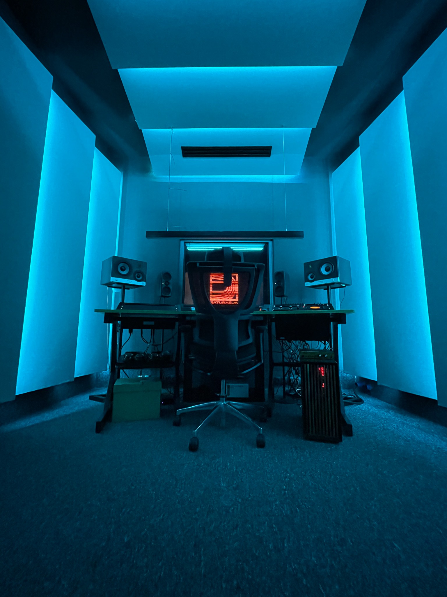 Profesjonalne studio nagrań z akustycznymi panelami ściennymi podświetlonymi na niebiesko, biurko z monitorem, głośnikami studyjnymi i sprzętem audio, krzesło biurowe na środku, oświetlenie ledowe...