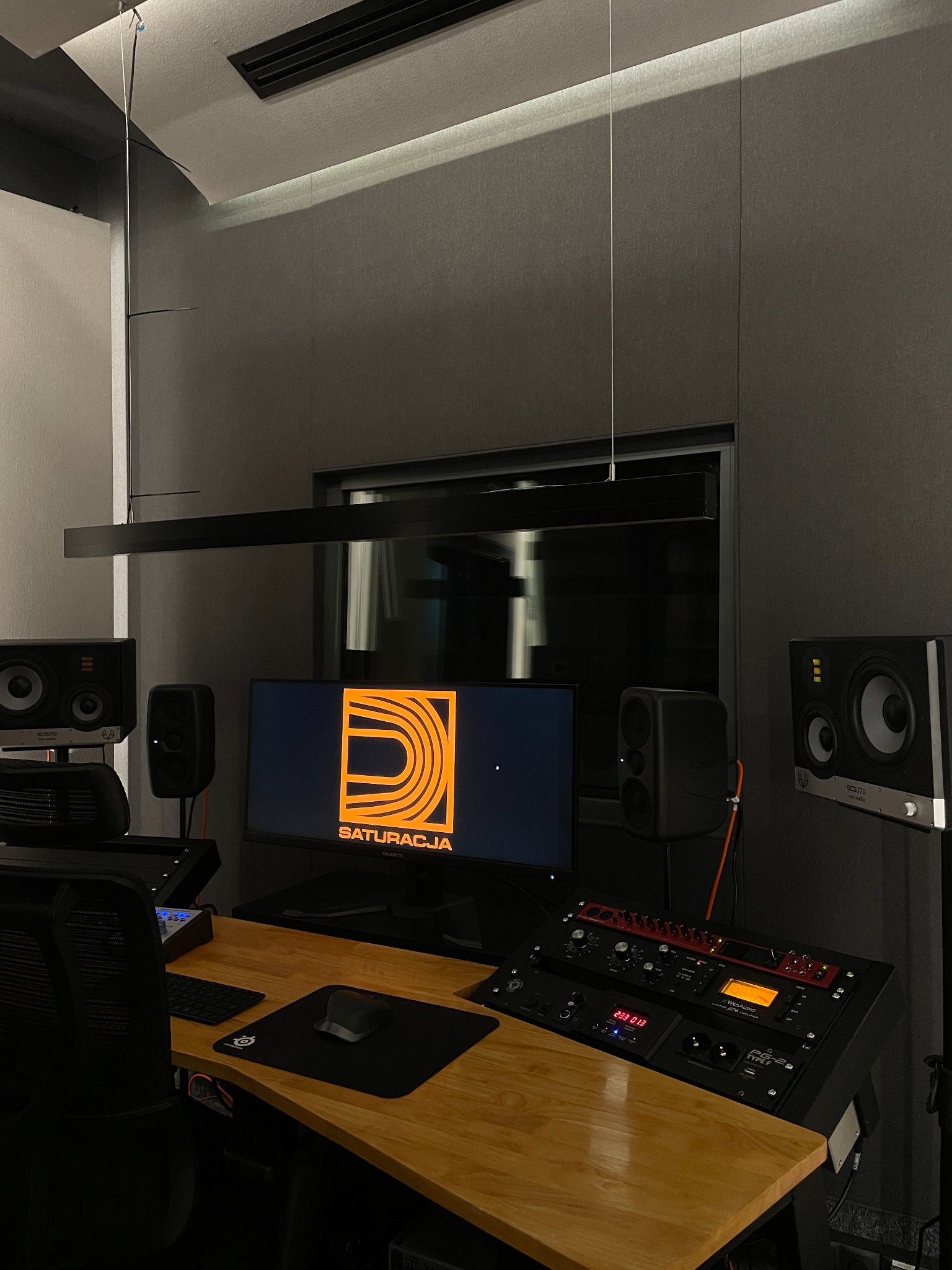 Wnętrze studia nagrań z drewnianym biurkiem, sprzętem audio, monitorami studyjnymi i ekranem z logo firmy 'Saturacja' w pomarańczowej kolorystyce.