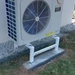 Biała pompa ciepła marki Rotenso zamontowana na białym stelażu na betonowej podstawie przy ścianie budynku z szarym kamieniem ozdobnym.