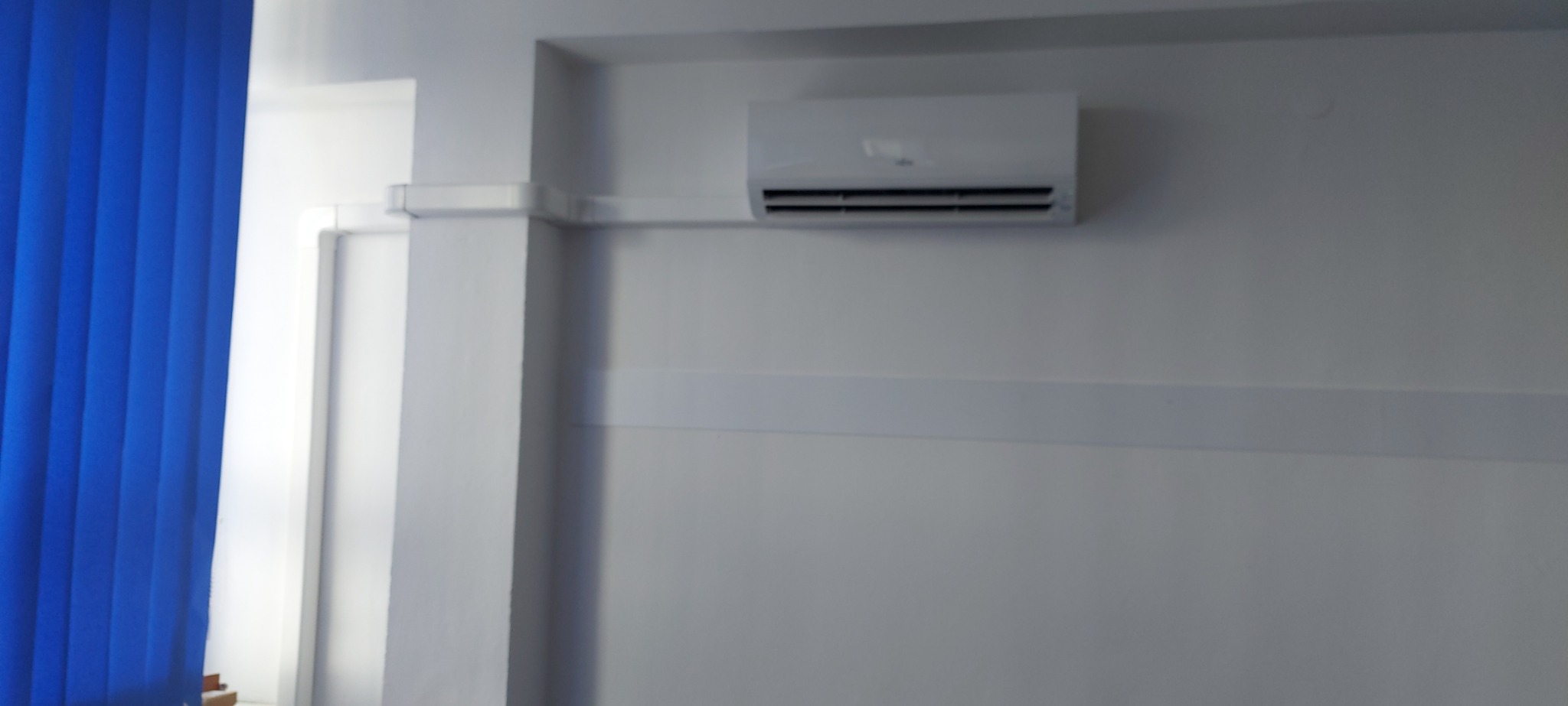 Biała jednostka wewnętrzna klimatyzacji zamontowana na ścianie biura, widoczne niebieskie żaluzje okienne po lewej stronie.