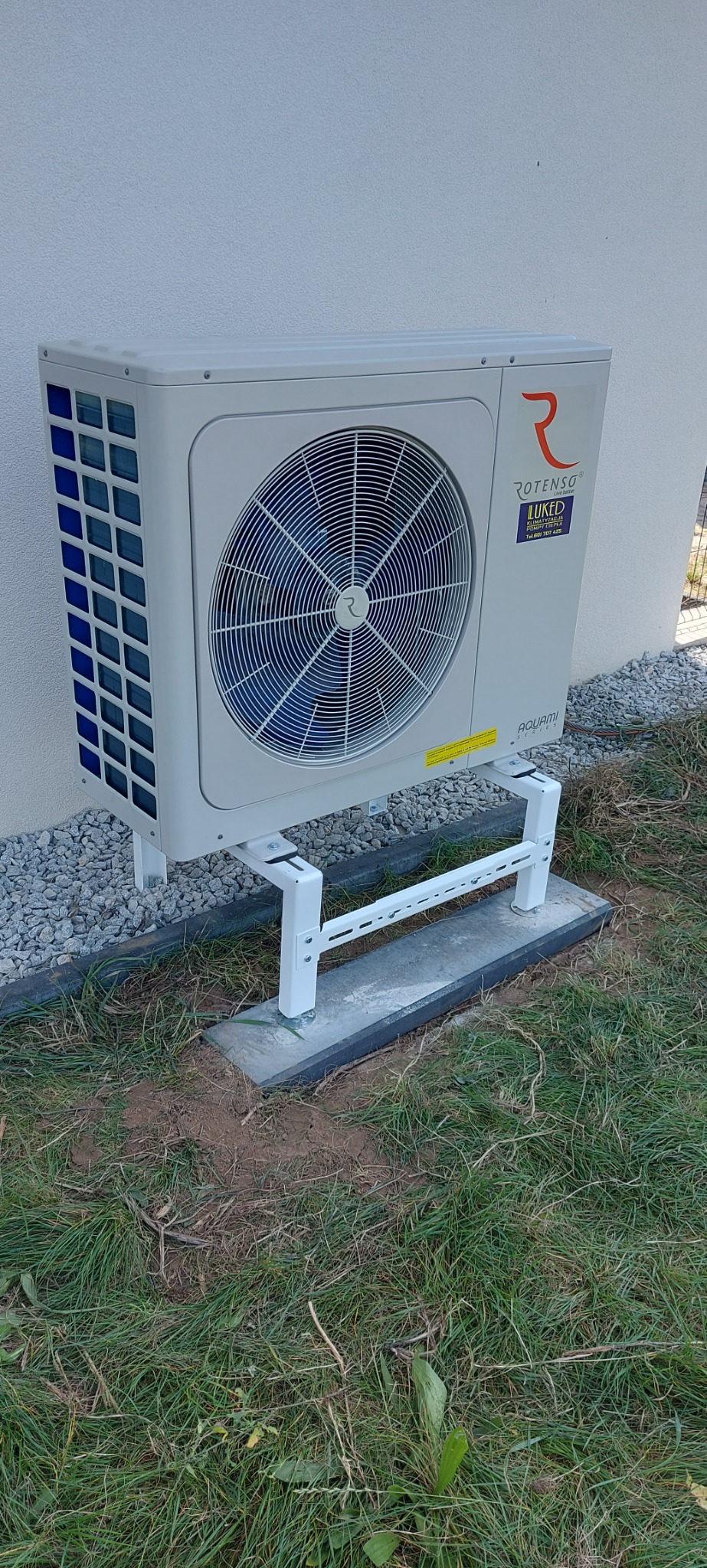 Biała pompa ciepła marki Rotenso zamontowana na białym stelażu na betonowej podstawie przy ścianie budynku z szarym kamieniem ozdobnym.