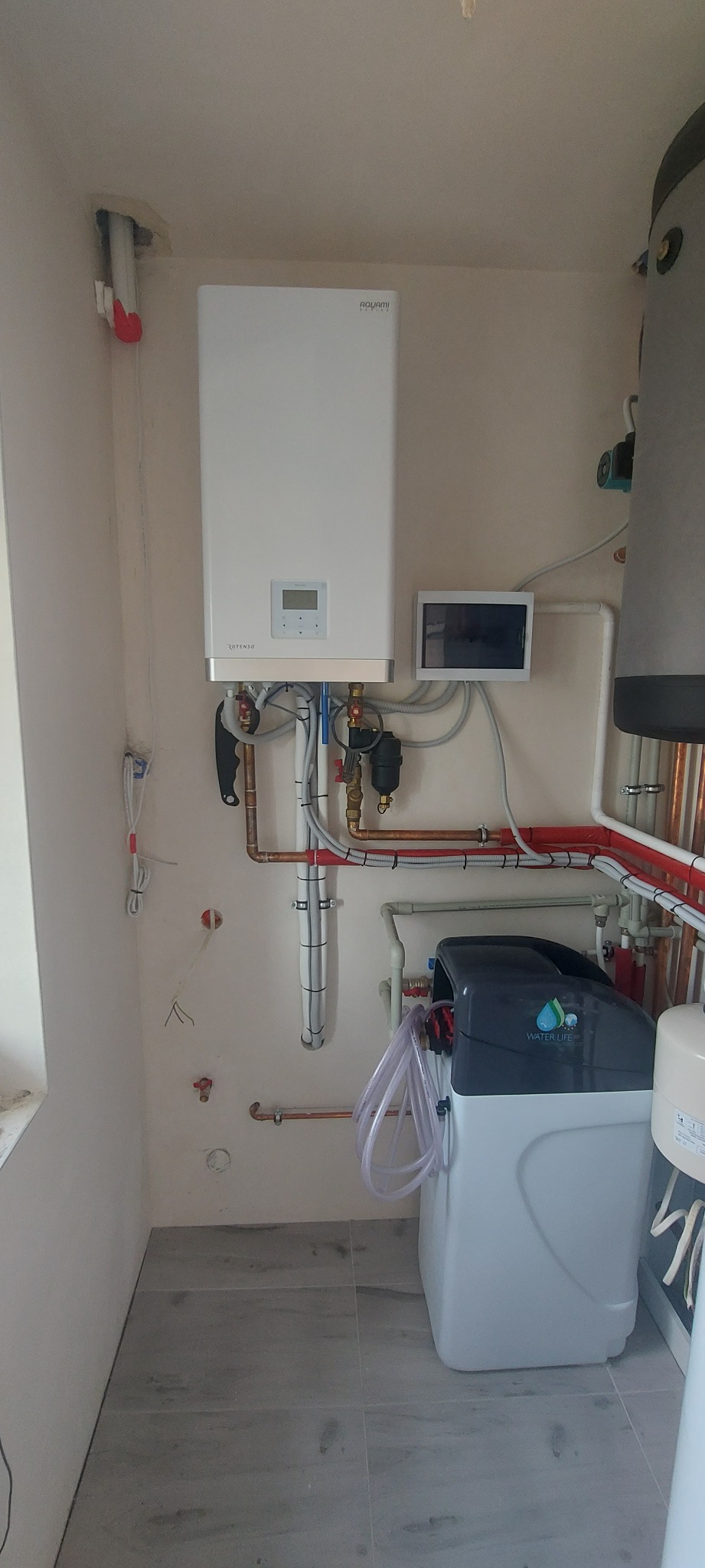 Wnętrze kotłowni z zamontowanym białym kotłem gazowym marki Rotenso, obok widoczny zmiękczacz wody Water Life oraz zasobnik ciepłej wody użytkowej, liczne rury miedziane i plastikowe z różnymi...