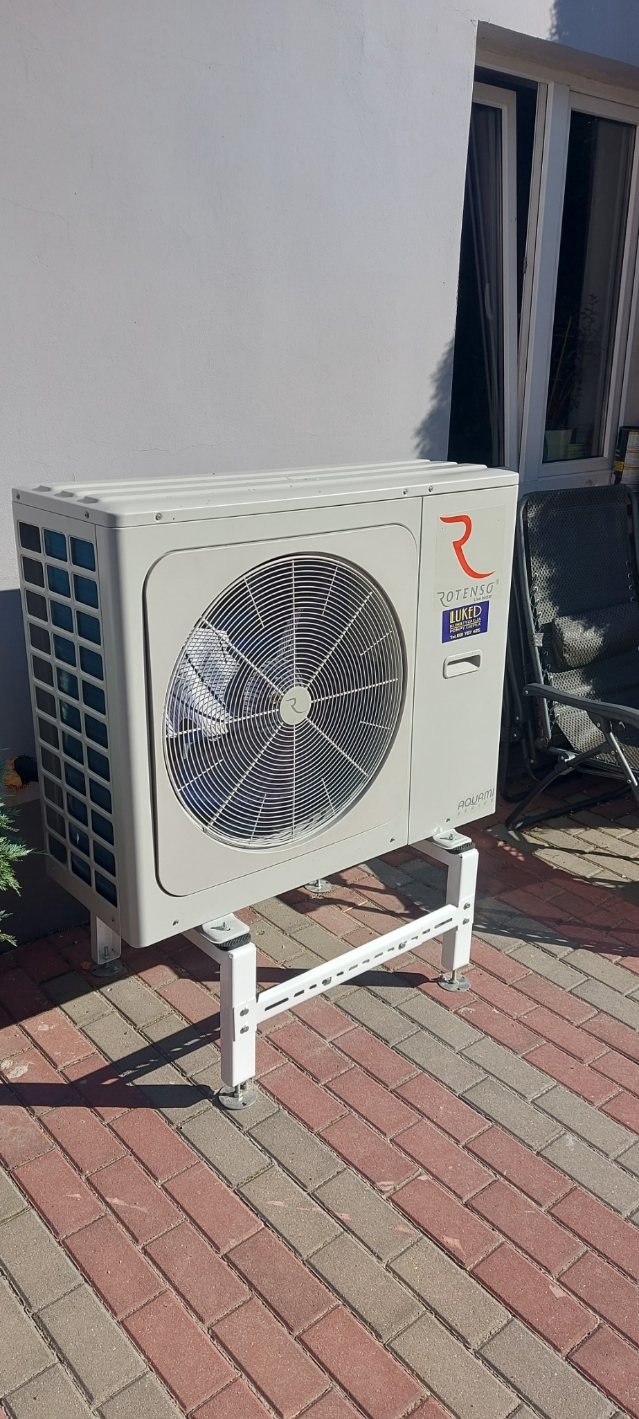 Zewnętrzna jednostka urządzenia grzewczego Rotenso Aquami Series zamontowana na białym stelażu, ustawiona na kostce brukowej przed białą ścianą budynku z oknem.