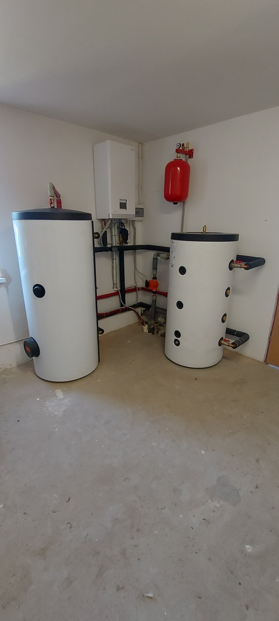 Dwa białe zbiorniki buforowe ciepła z czarnymi pokrywami i przyłączami, instalacja grzewcza z kotłem gazowym i czerwonym naczyniem wzbiorczym na ścianie w pomieszczeniu technicznym z surową podłogą.