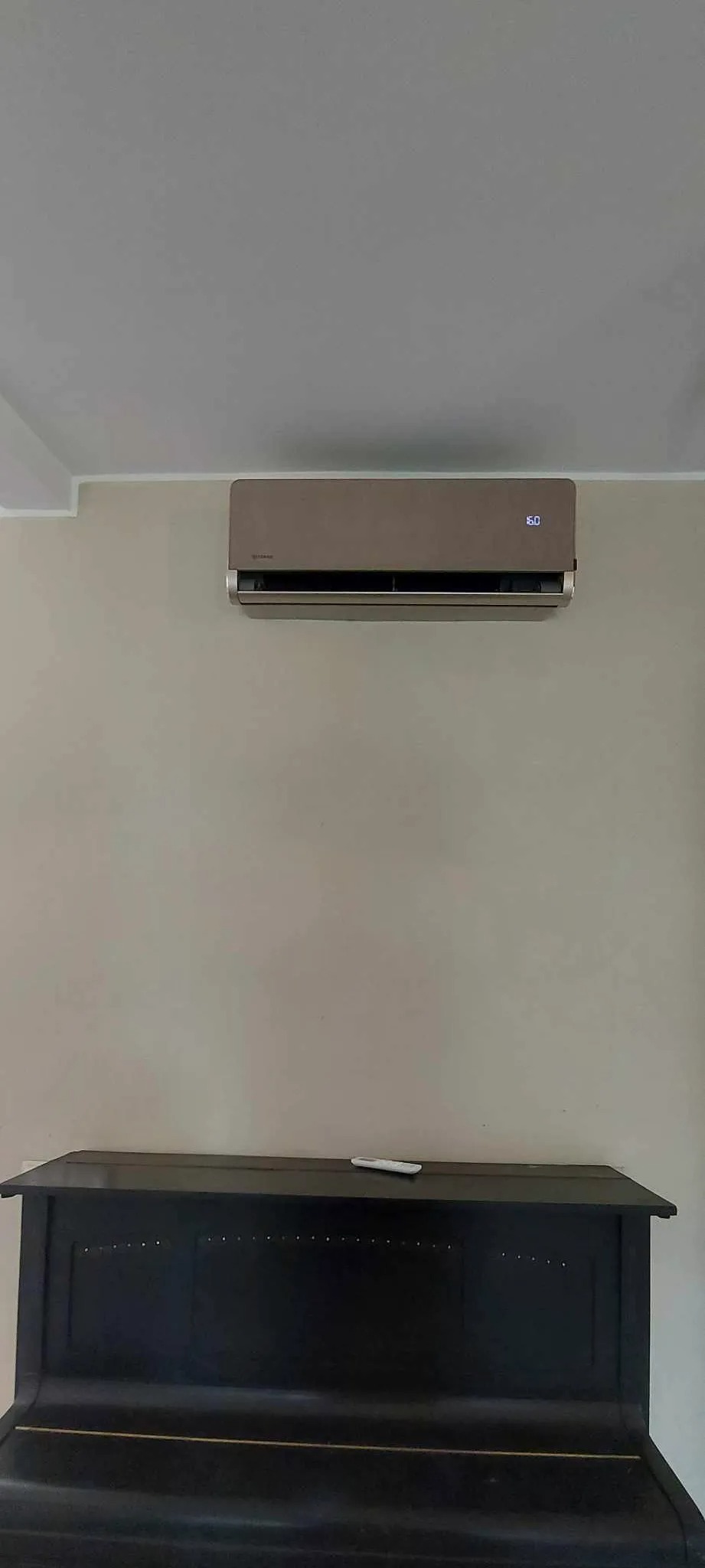 Beżowa klimatyzacja ścienna z wyświetlaczem temperatury 16 stopni Celsjusza, zamontowana nad czarnym pianinem w pokoju z beżowymi ścianami.