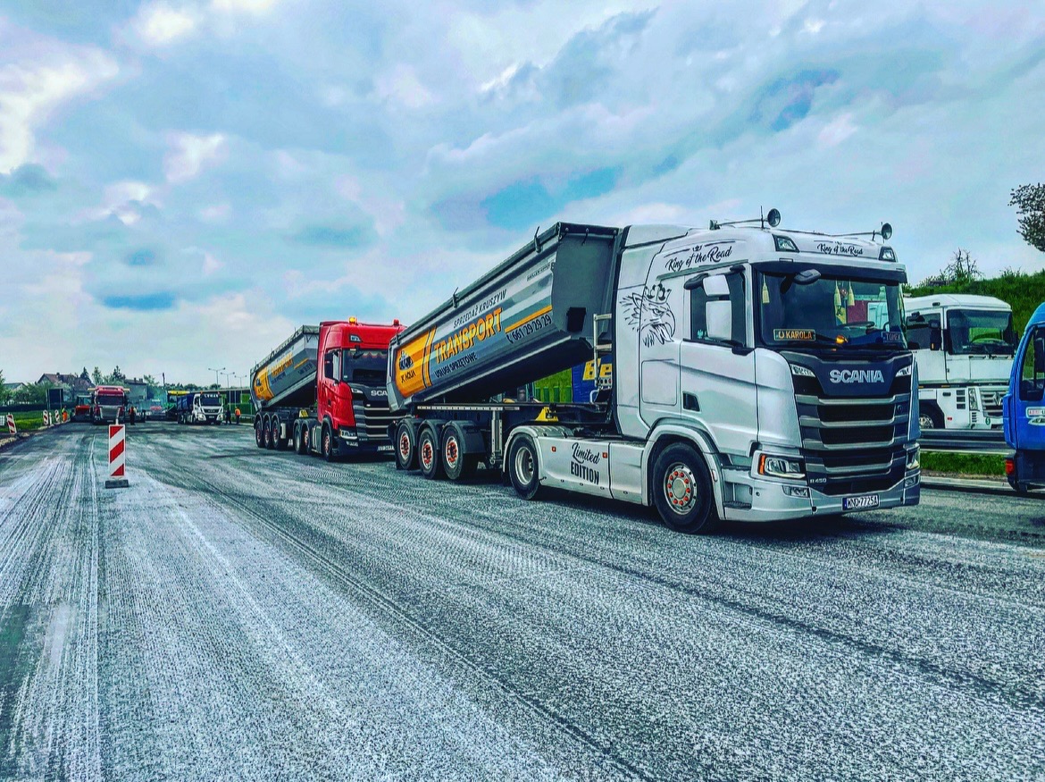 Dwa samochody ciężarowe Scania z naczepami wywrotkami, jeden biały z grafiką wilka i napisem 'Limited Edition', drugi czerwony, stoją na nowo położonej, szarej nawierzchni. W tle widoczne inne...