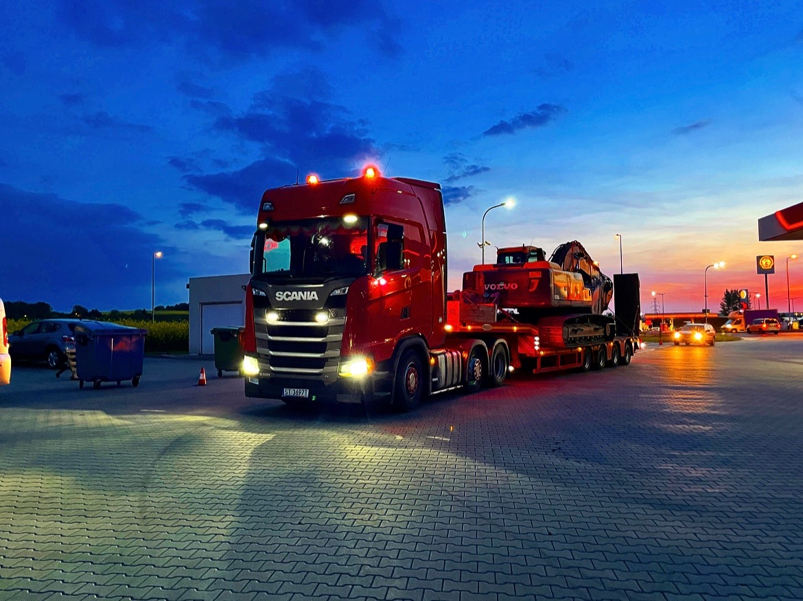 Czerwona ciężarówka Scania z naczepą niskopodwoziową przewożąca pomarańczową koparkę Volvo, sfotografowana na stacji benzynowej o zmierzchu, z widocznym niebem w odcieniach niebieskiego...