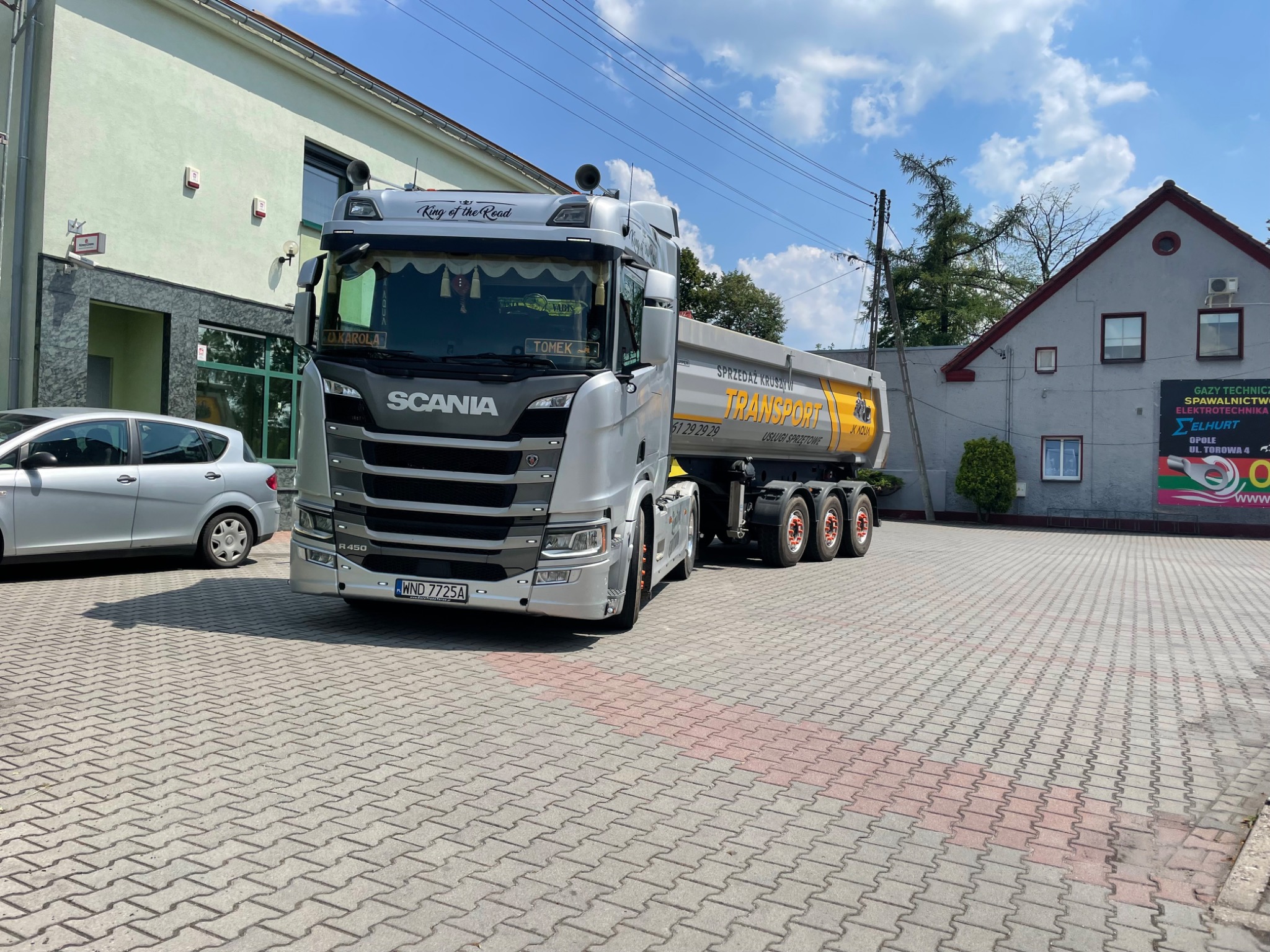 Srebrno-szara ciężarówka Scania z naczepą wywrotką, z napisem 'TRANSPORT' w kolorze żółtym, zaparkowana na placu wyłożonym kostką brukową, na tle budynku z reklamą i fragmentem zaparkowanego...