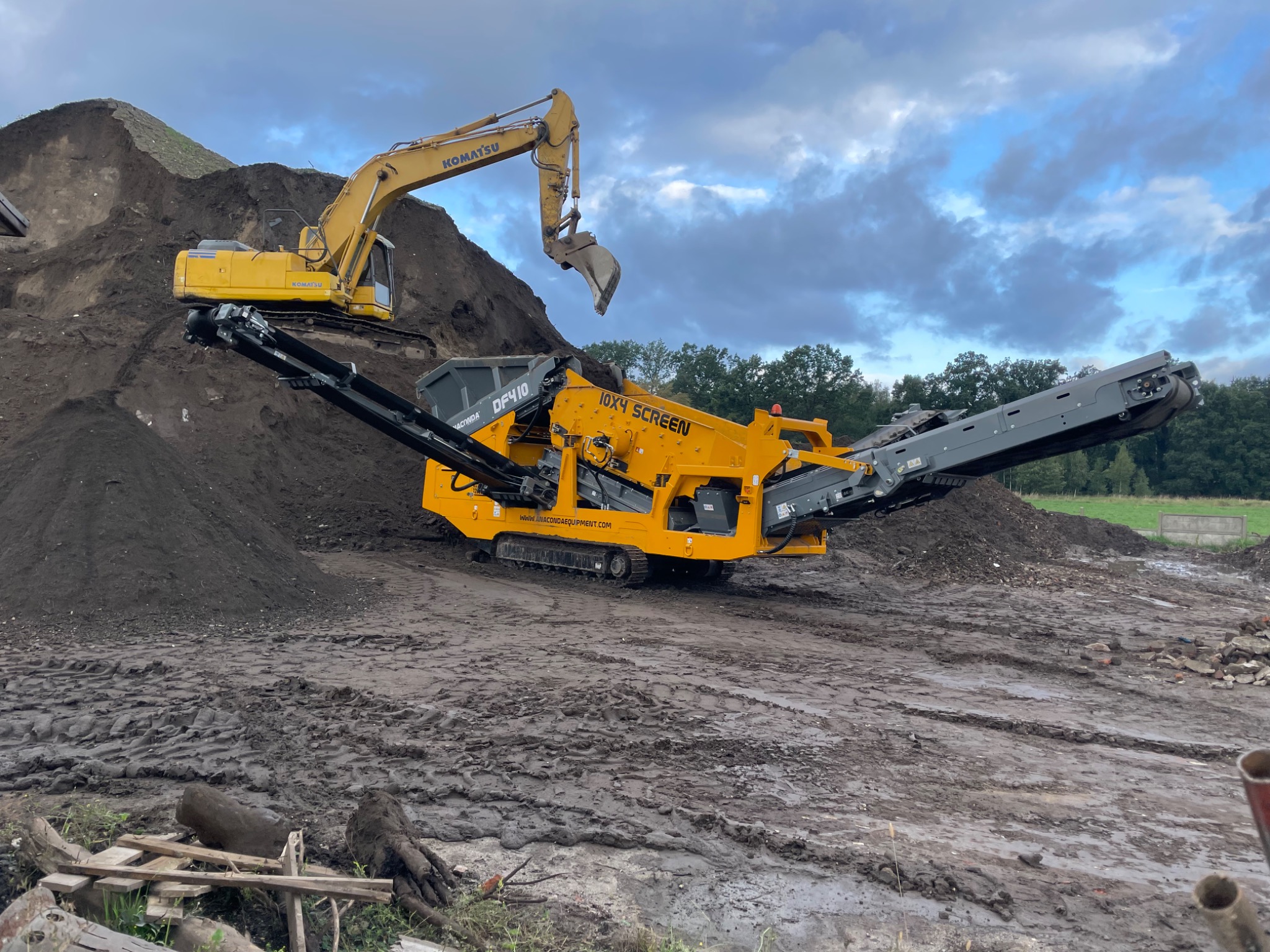 Żółty koparka Komatsu i przesiewacz mobilny Anaconda DF410 na placu budowy z górą ziemi w tle, pochmurne niebo.