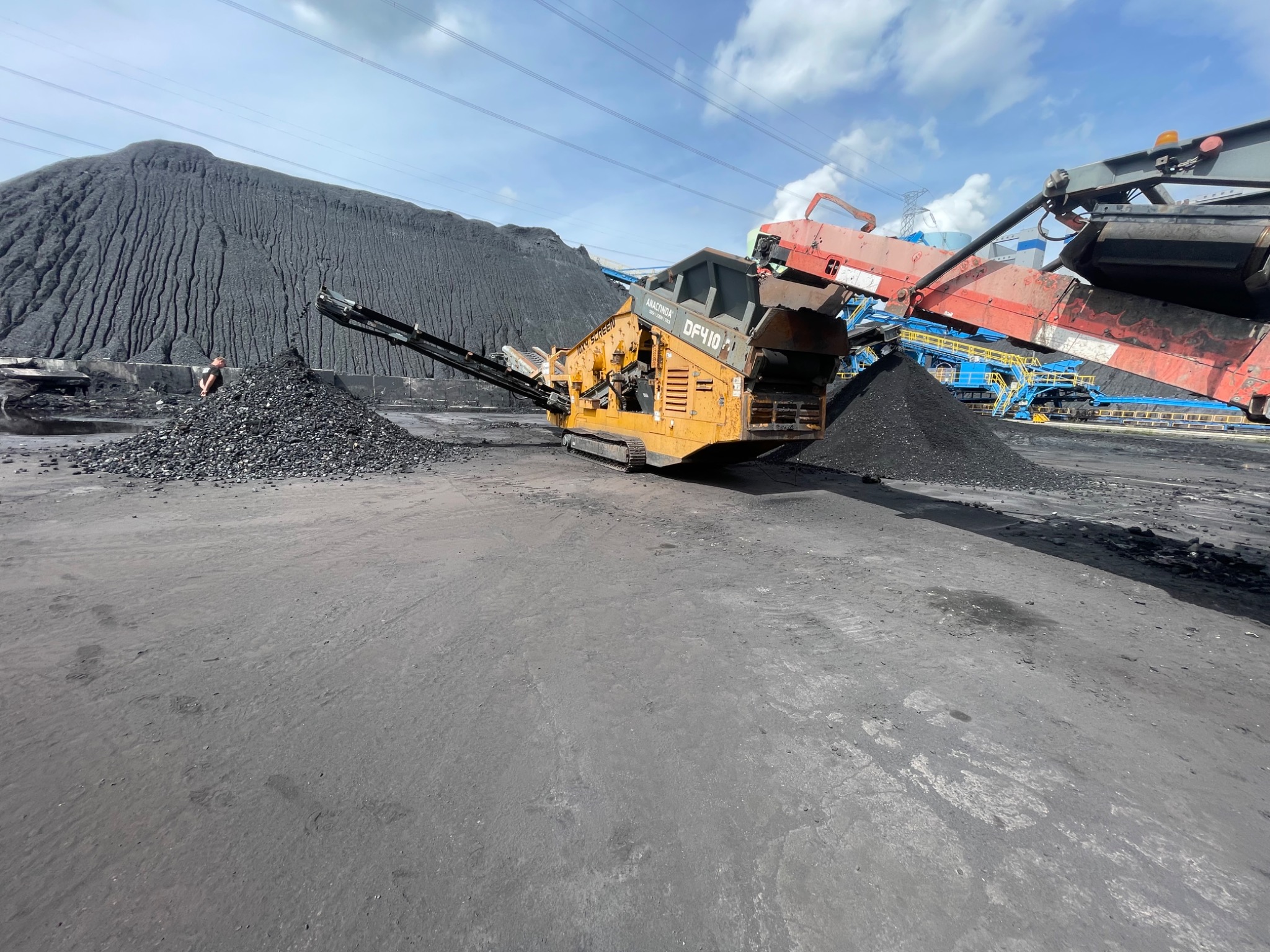 Żółty mobilny przesiewacz do kruszyw z podajnikiem, pracujący przy hałdzie ciemnego materiału sypkiego, prawdopodobnie węgla, z widocznymi elementami innych maszyn i fragmentem sylwetki osoby w tle.