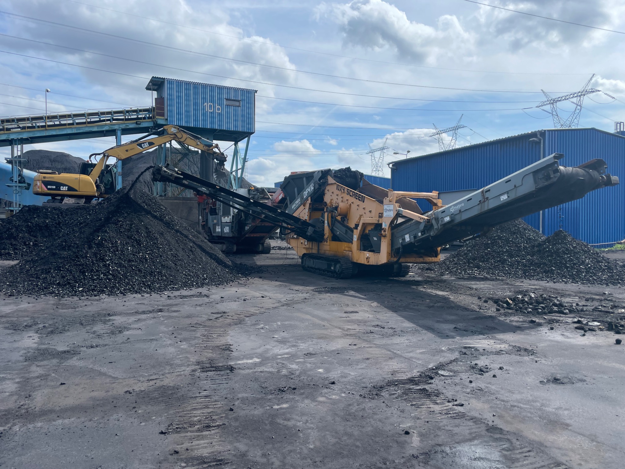 Żółty koparka CAT wysypuje czarny węgiel na przesiewacz mobilny Powerscreen, w tle niebieski budynek przemysłowy i słupy wysokiego napięcia.