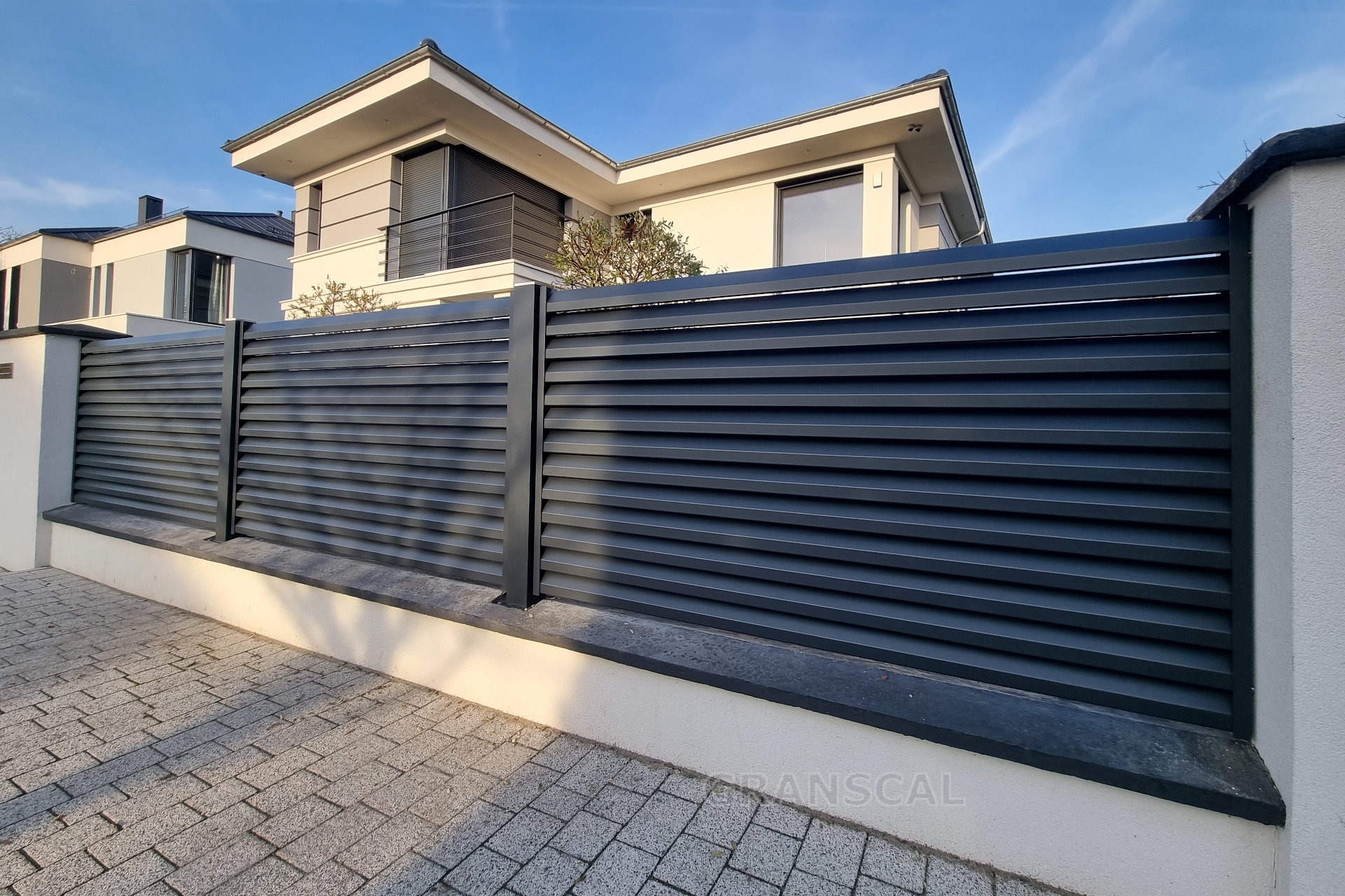 Nowoczesne, poziome, grafitowe ogrodzenie panelowe na niskiej, białej podmurówce, z widocznym fragmentem kostki brukowej i domu w tle, oświetlone popołudniowym słońcem.