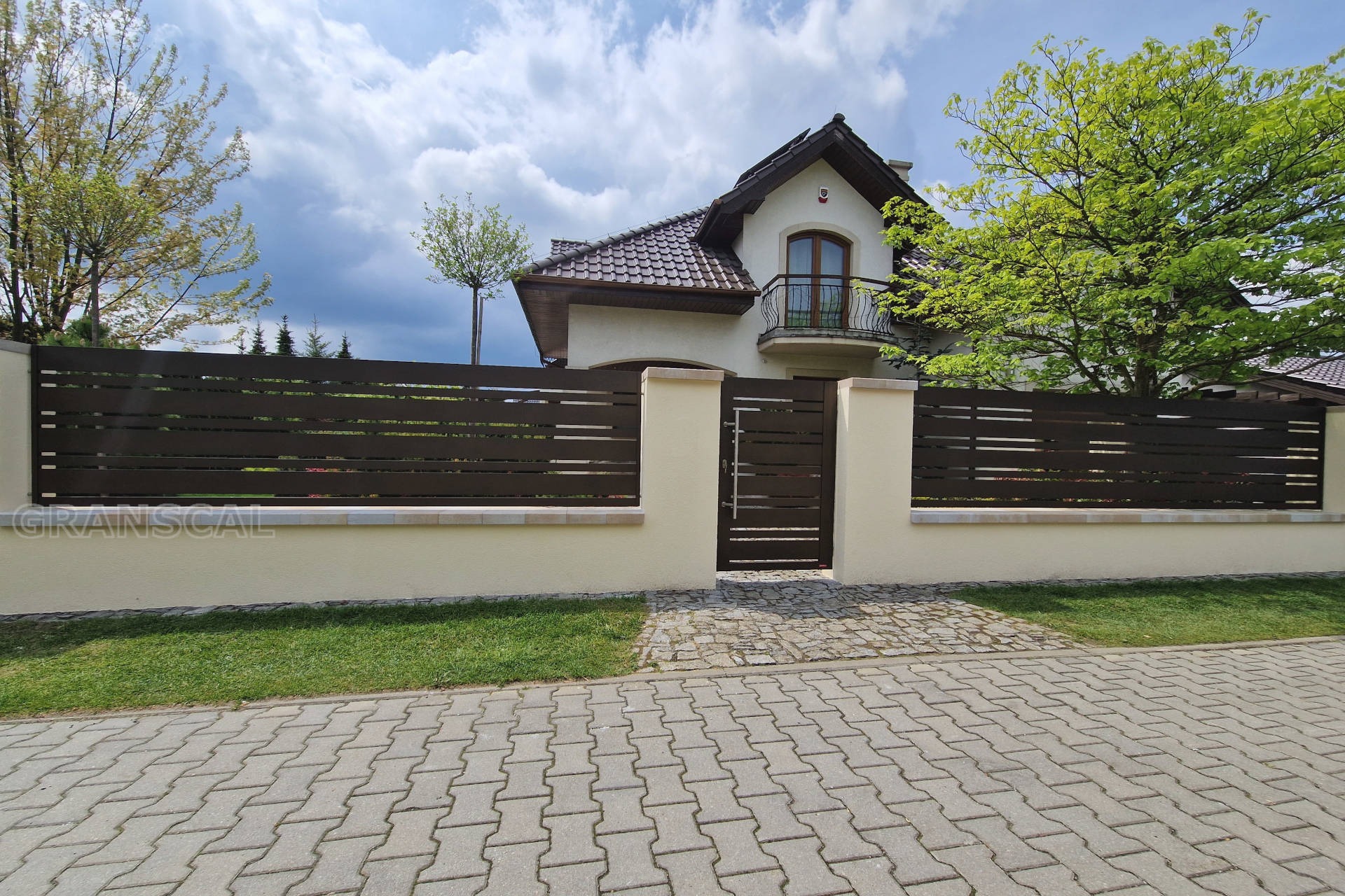 Nowoczesne, poziome ogrodzenie panelowe w kolorze ciemnego brązu, osadzone na niskim, beżowym murku z filarami, z bramką wejściową, na tle domu z balkonem i zielenią drzew.