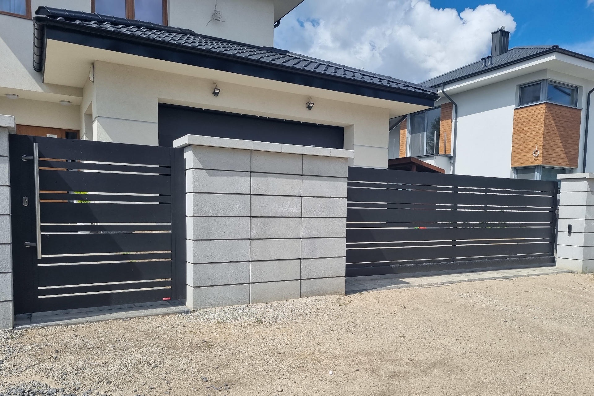 Nowoczesna, pozioma brama wjazdowa i furtka z czarnych profili, osadzona na betonowych słupkach z szarej kostki, na tle domu z ciemnym dachem i jasną elewacją.