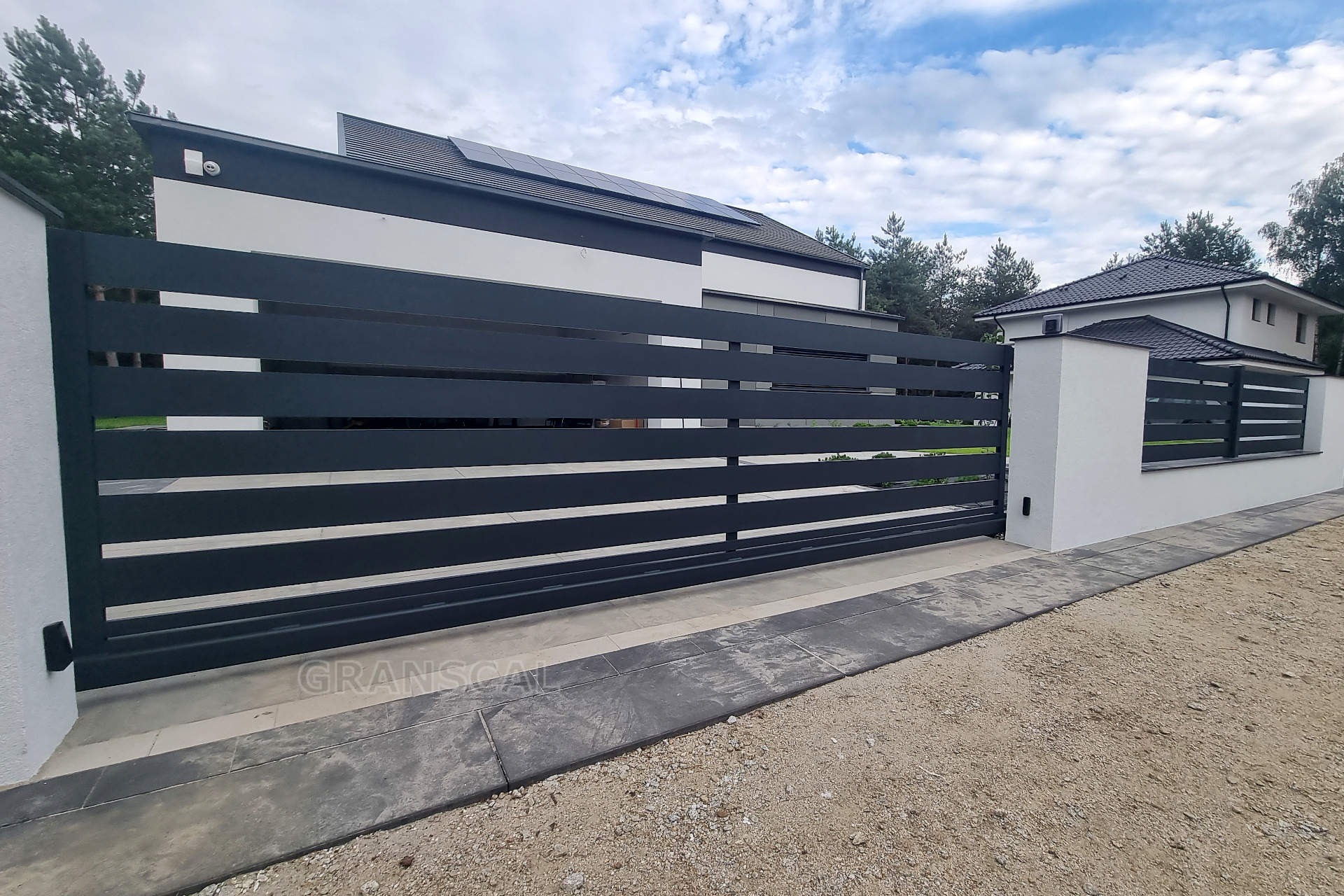 Nowoczesna, przesuwna brama w kolorze grafitowym z poziomymi panelami, zamontowana przed domem z białą elewacją i szarym dachem, widoczna kostka brukowa i żwir.