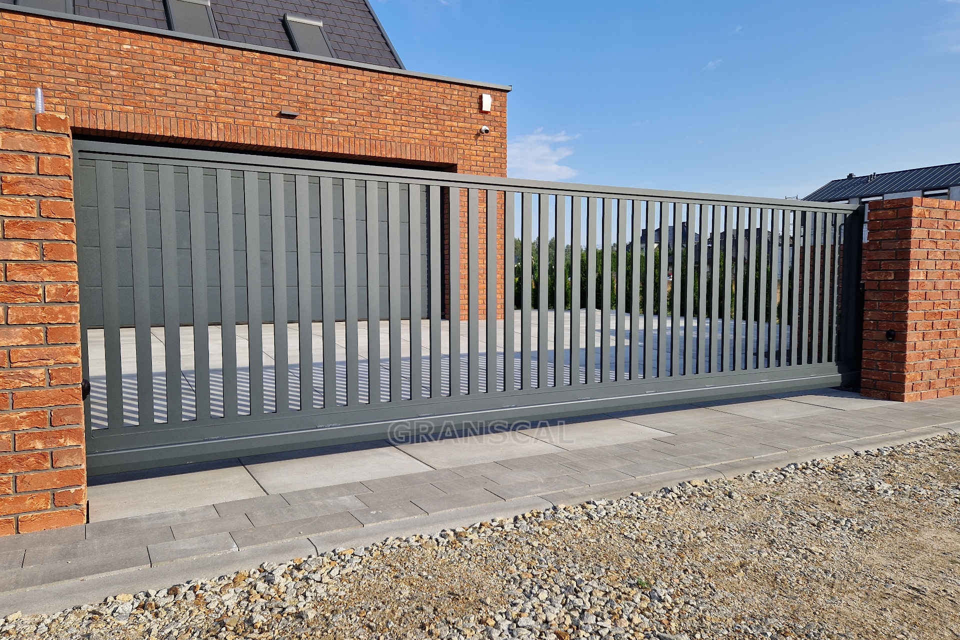 Nowoczesna, przesuwna brama w kolorze grafitowym z pionowymi panelami, zamontowana w Zakrzewie, na tle ceglanego domu z grafitowym dachem i błękitnym niebem.