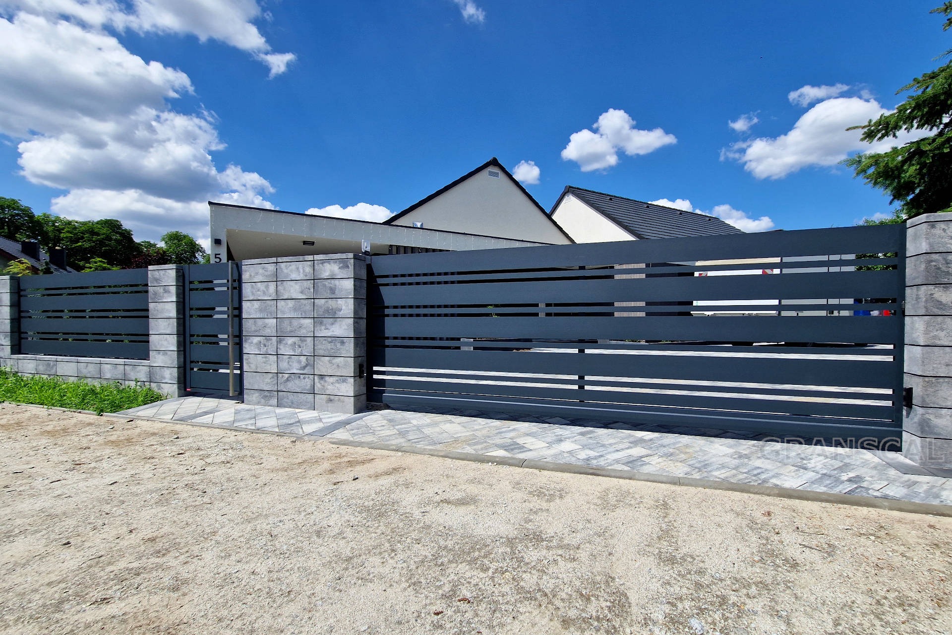 Nowoczesne, poziome, metalowe ogrodzenie w kolorze grafitowym z przesuwną bramą, wykończone szarymi, betonowymi słupkami i podmurówką, prowadzące do posesji z domem o białej elewacji pod błękitnym...