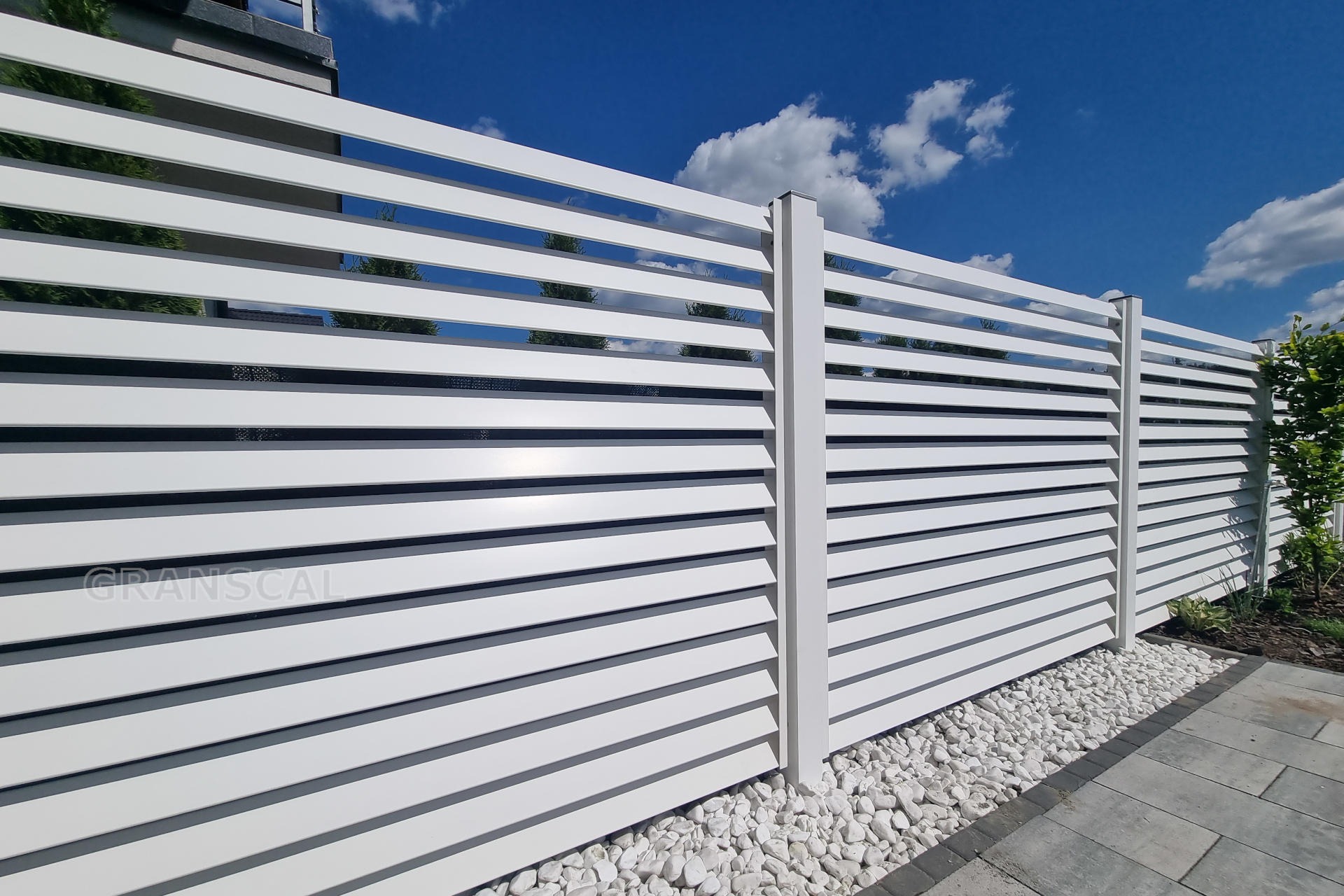 Nowoczesne, białe ogrodzenie panelowe z poziomych listew, uzupełnione białym kamieniem ozdobnym u podstawy, na tle błękitnego nieba z chmurami.