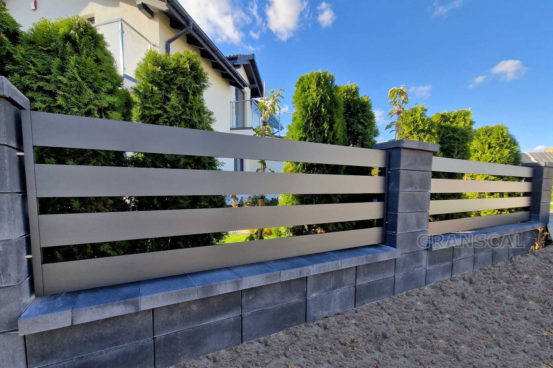 Nowoczesne ogrodzenie panelowe w kolorze szarym z poziomymi szczeblami, osadzone na murku z szarych bloczków, z widocznym domem i tujami w tle oraz logo 'GRANSCAL' na jednym ze słupków.