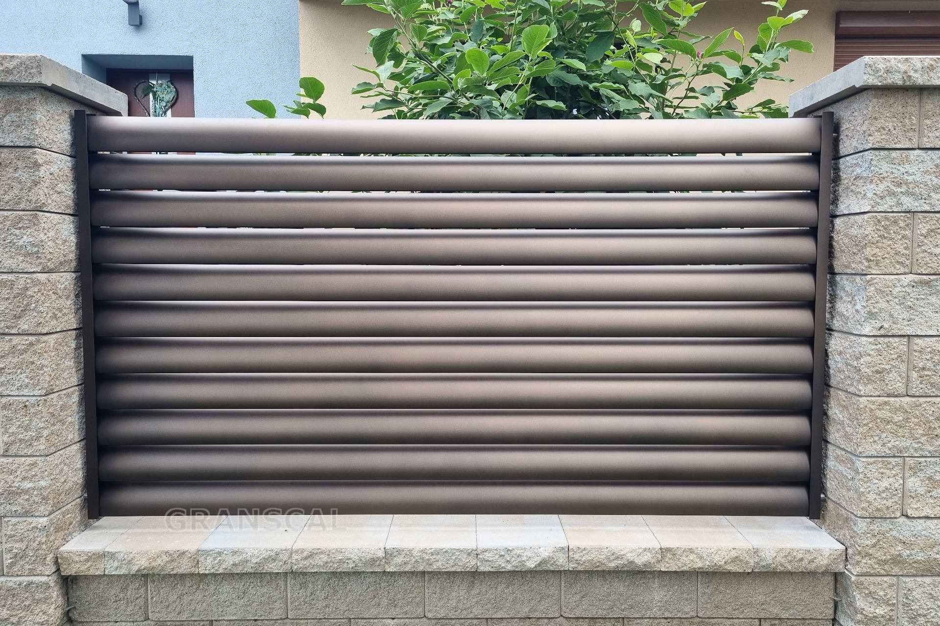 Nowoczesne ogrodzenie lamelowe w kolorze brązowym, z kamiennymi słupkami i podmurówką, widok z przodu na tle domu z zielenią.