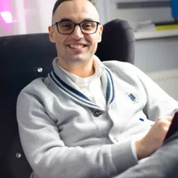 Uśmiechnięty mężczyzna w okularach siedzi w fotelu, trzyma długopis i notatnik, w tle biuro z kwiatami i dokumentami.