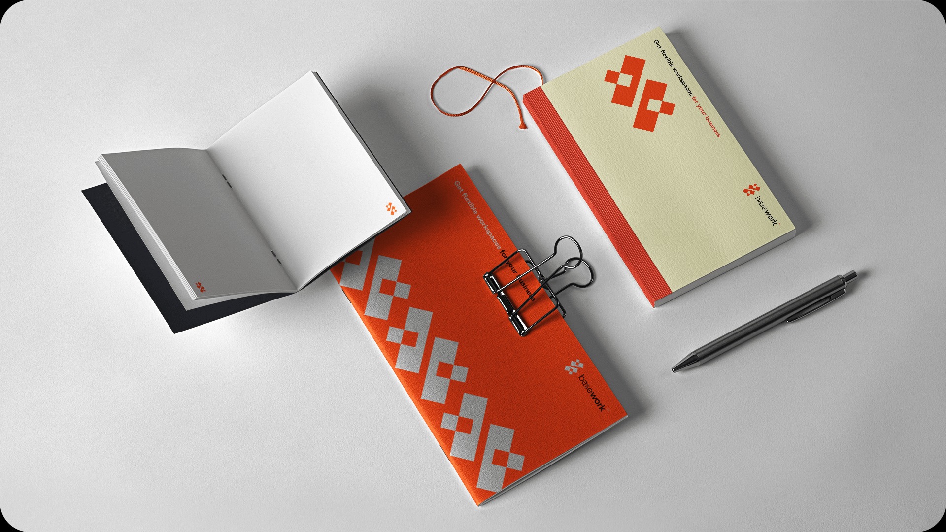 Aranżacja na białym tle: otwarty notatnik z czarną okładką, pomarańczowy i kremowy notes z logo, spinacz biurowy i długopis.