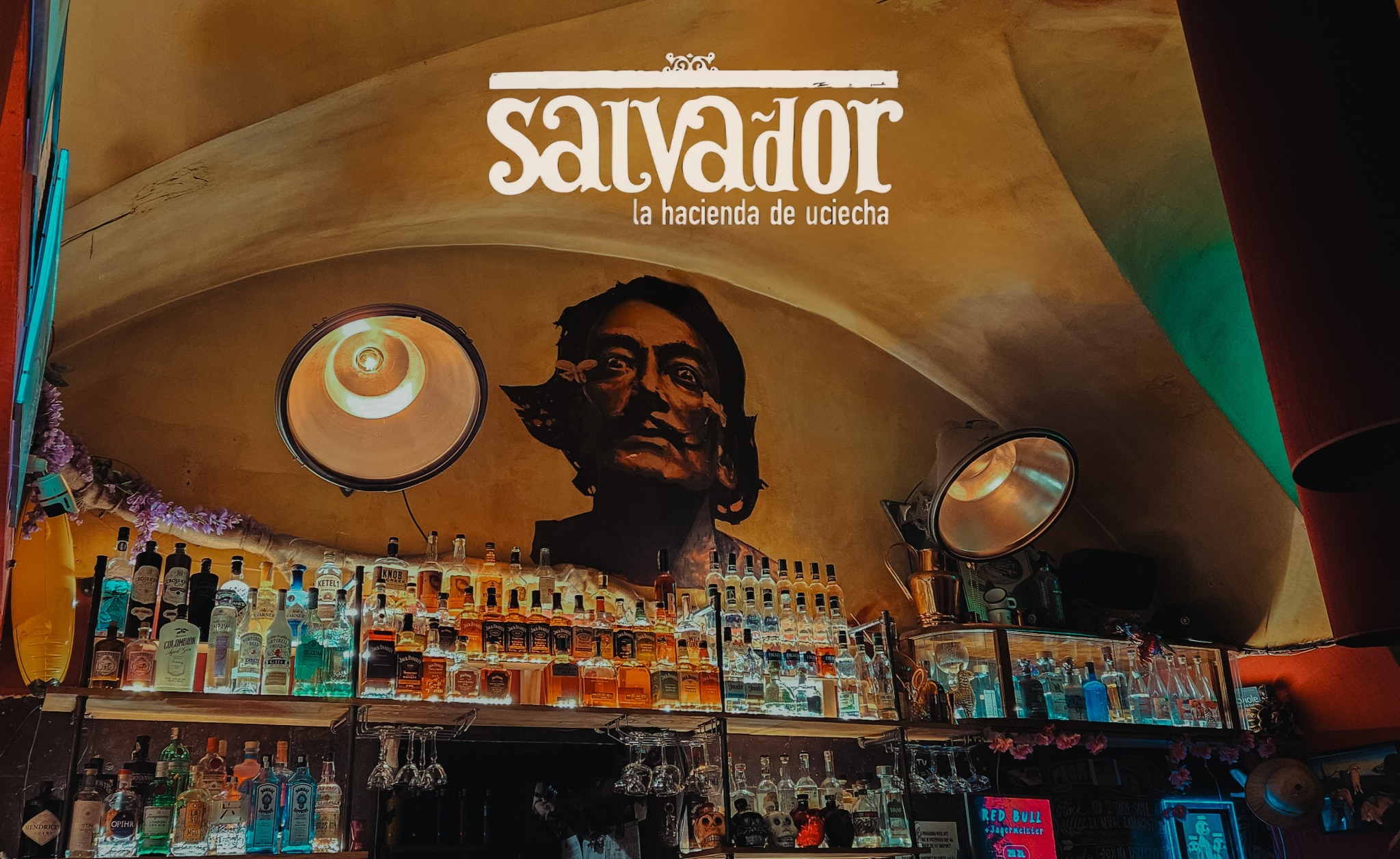 Bar z półkami pełnymi butelek alkoholu, oświetlony lampami, z muralem Salvadora Dali na ścianie. Lokal 'Salvador, la hacienda de uciecha'.