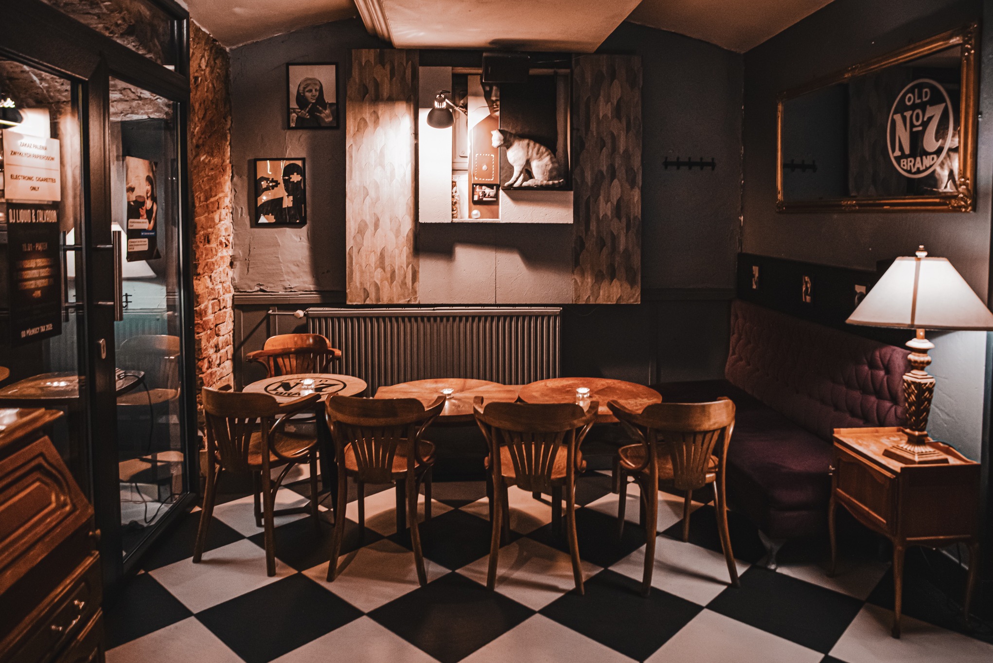 Klimatyczne wnętrze baru 'Loża Kot' z drewnianymi krzesłami przy okrągłych stolikach, czarno-biała podłoga i dekoracje, w tym obraz z kotem. Nastrojowe oświetlenie.